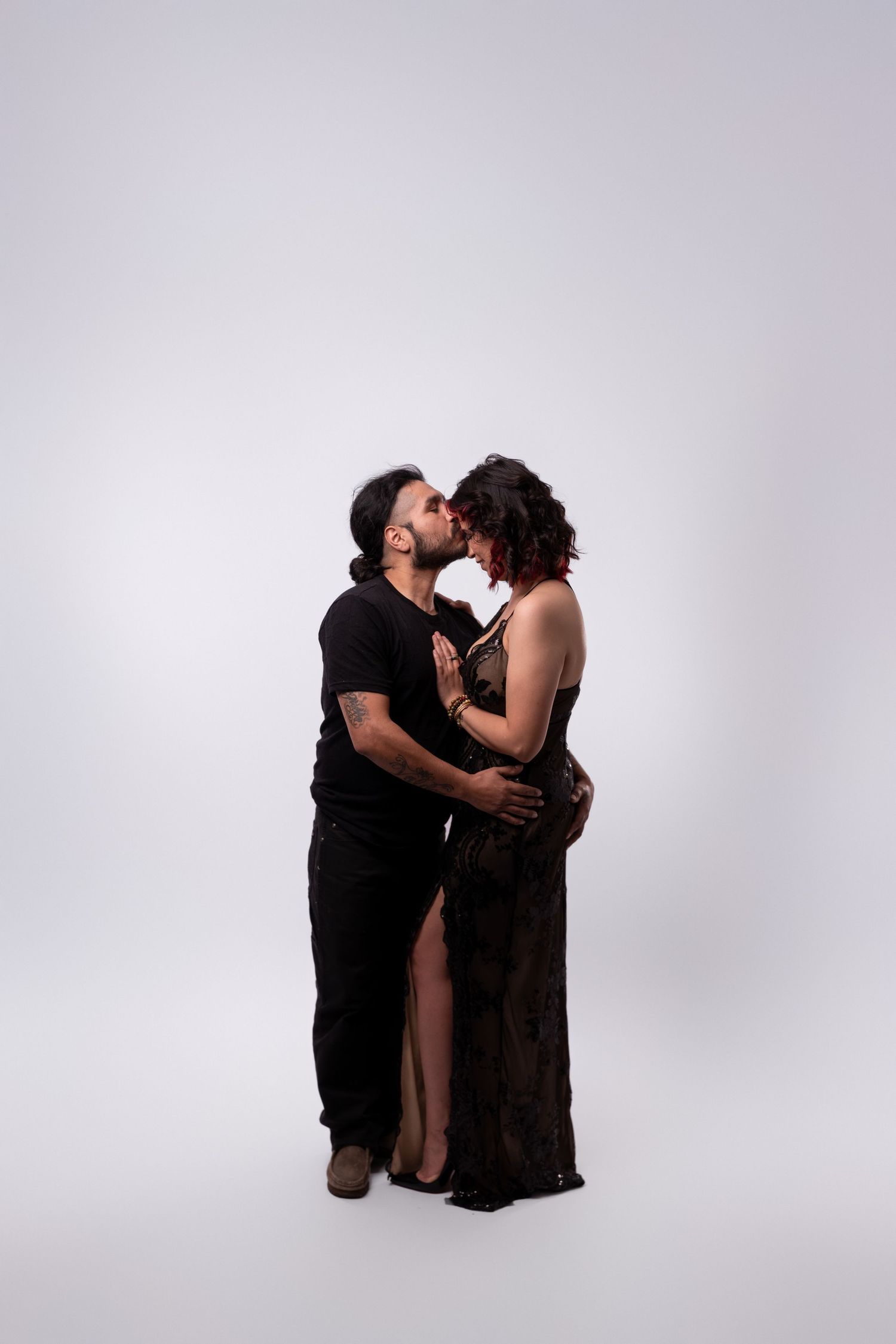 Couple in black attire embracing against white backdrop in studio setting.