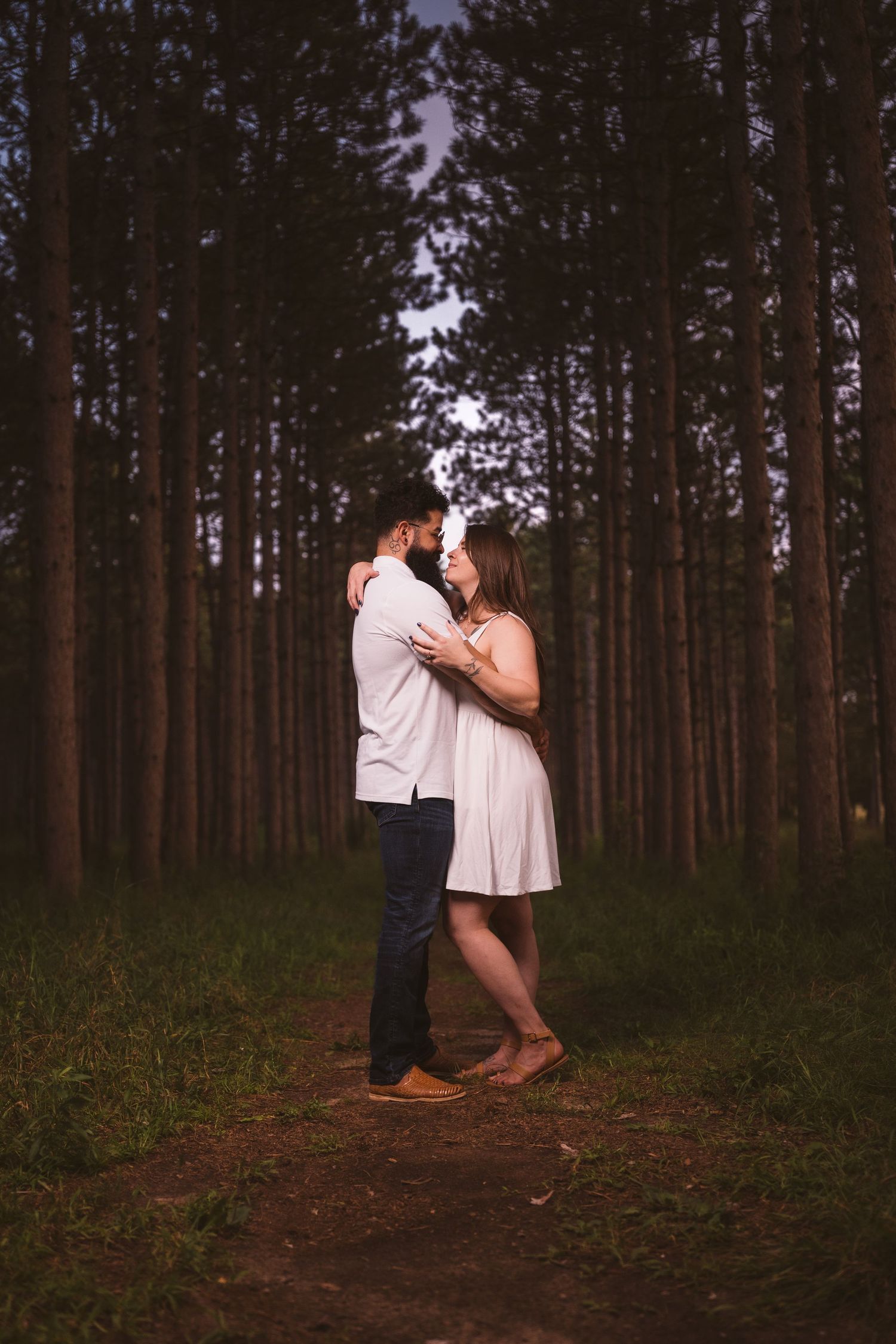A couple shares an intimate moment on a forest trail surrounded by towering pine trees at dusk.