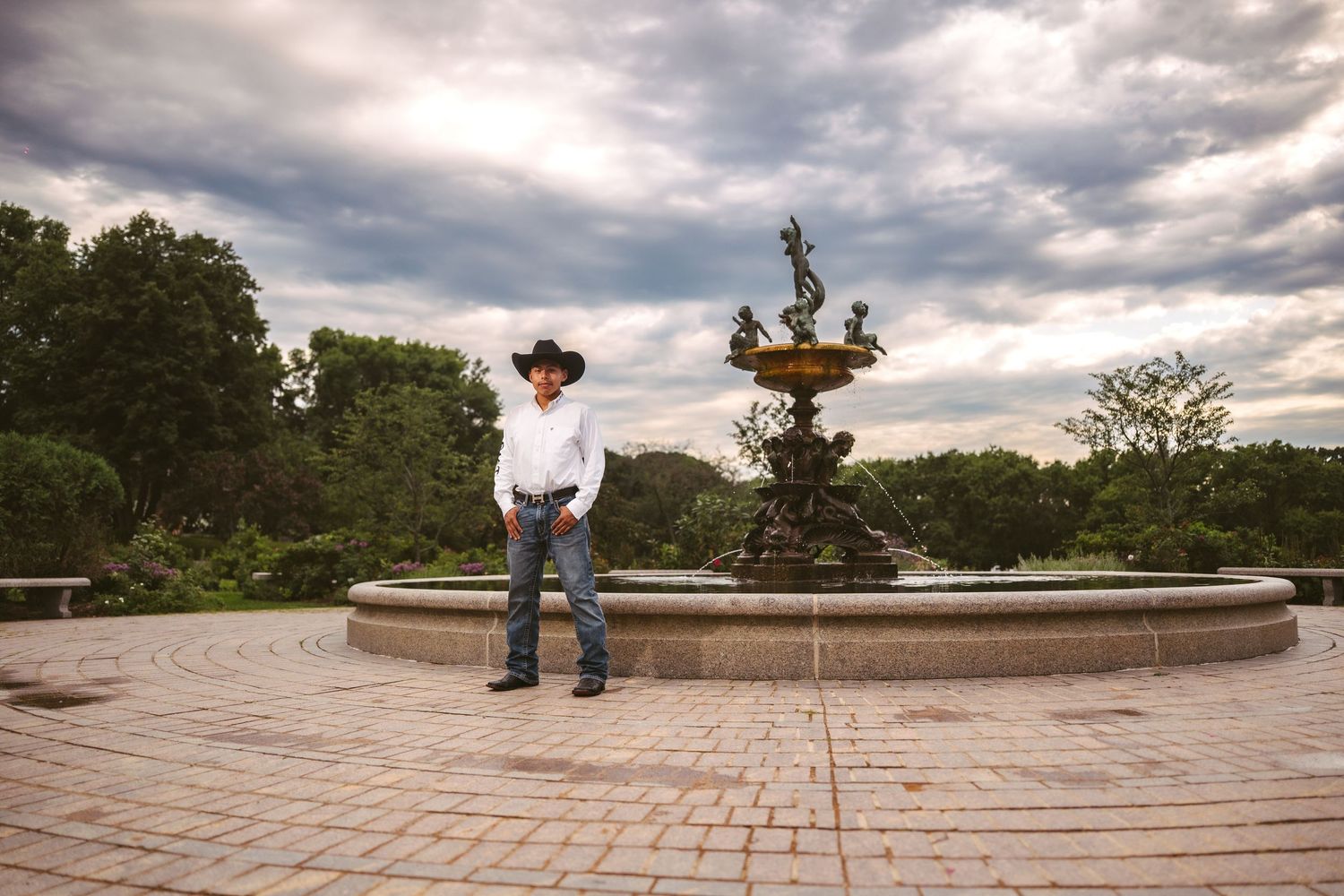 Dramatic cloudy sky over a circular brick patio with ornate fountain and statues at sunset.