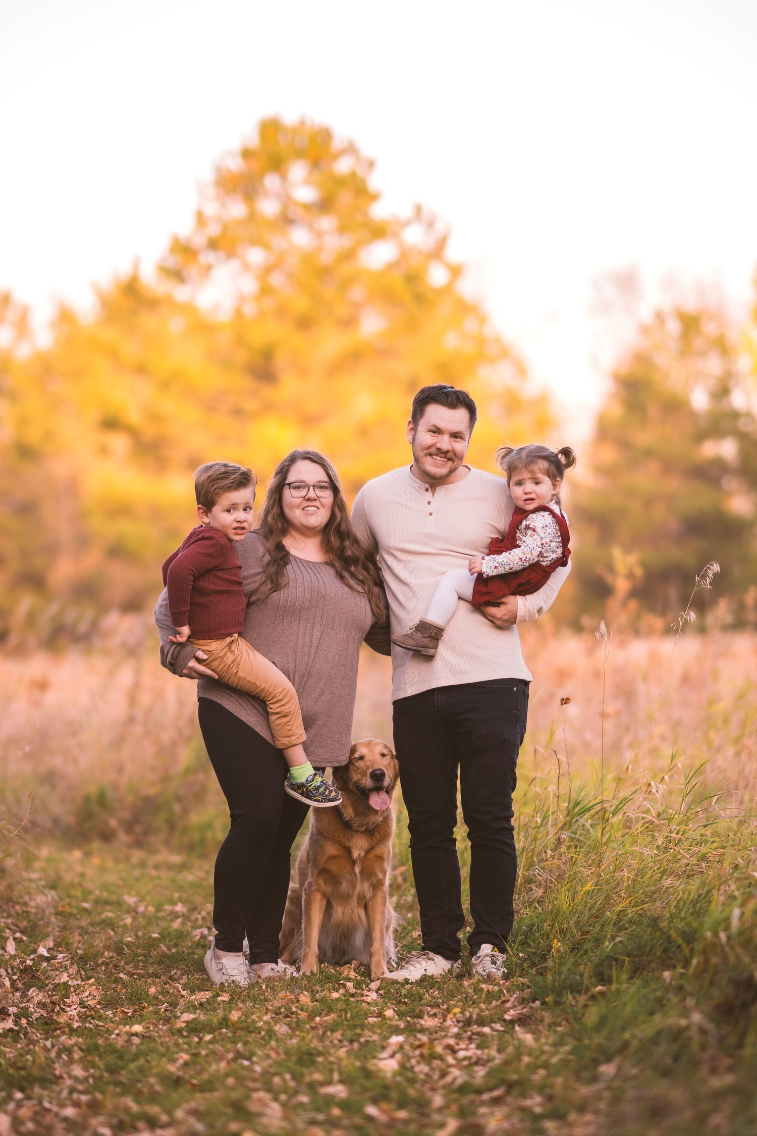 Family and dog pose together in field with golden fall foliage.