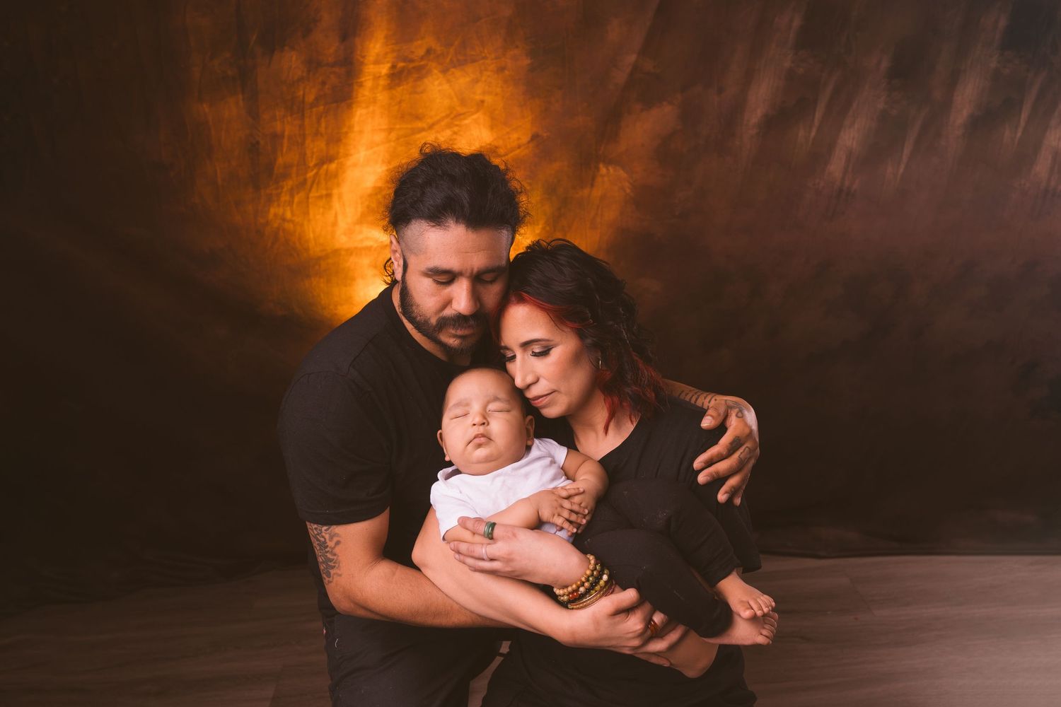 Family embracing on wooden floor with warm lighting and dark backdrop.