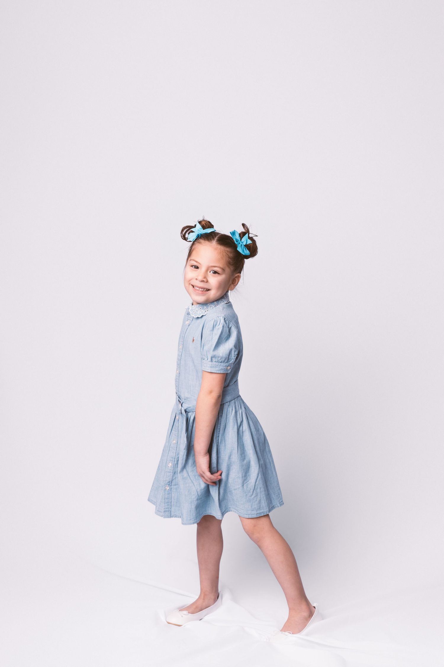 A toddler in a light blue dress with pigtails twirls and plays against a white studio backdrop.