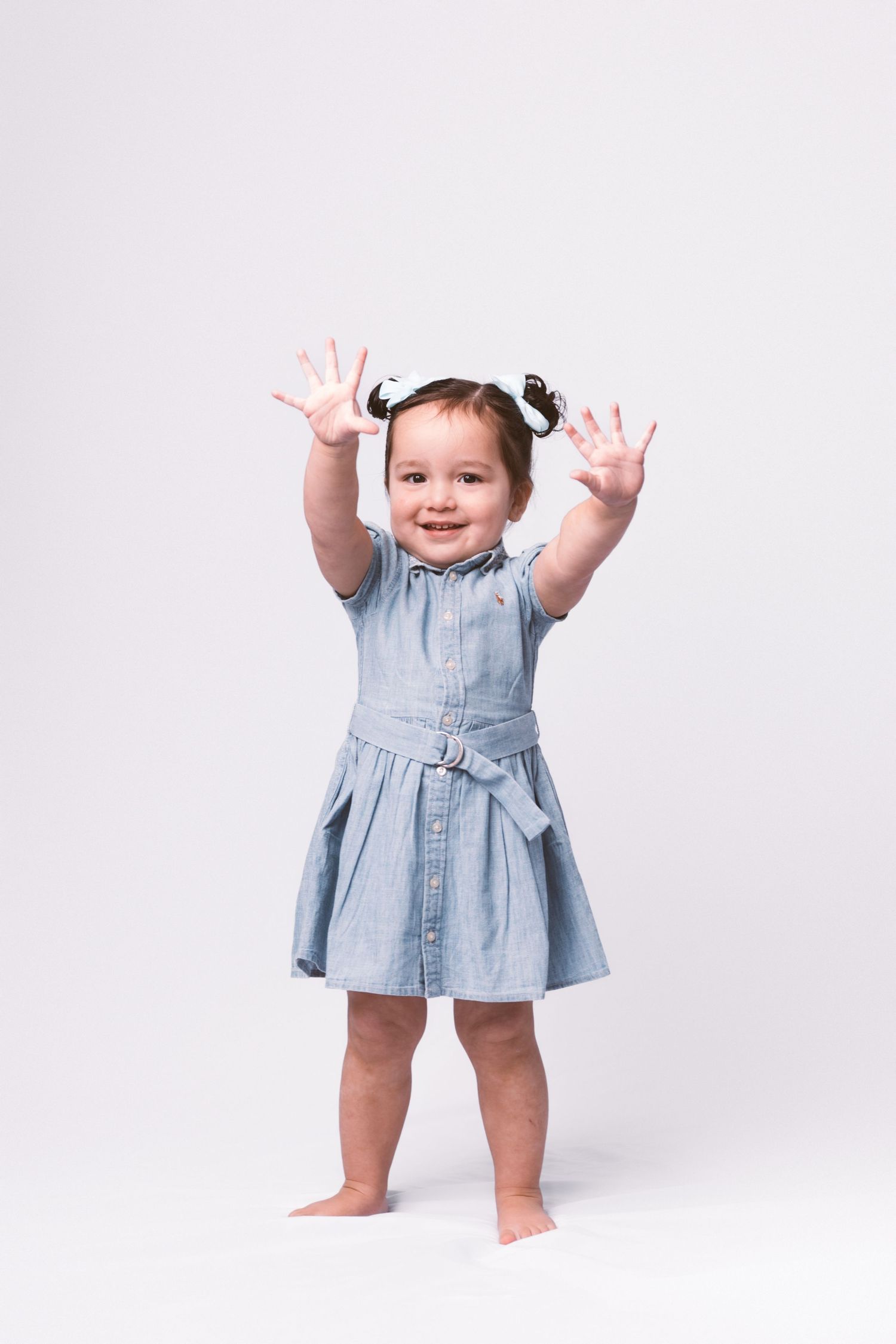 A toddler in a denim dress raises their hands playfully while standing against a white backdrop.