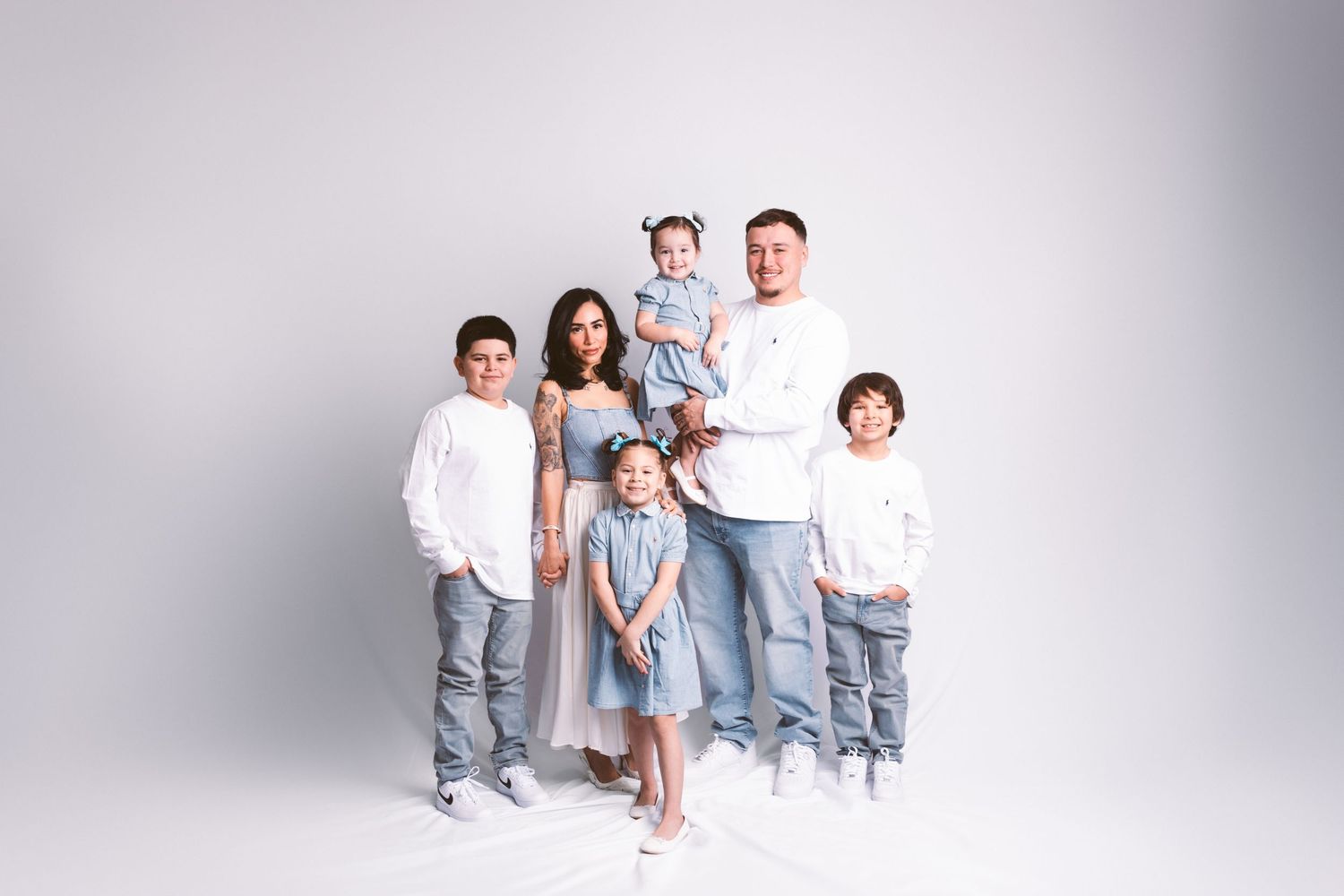 Family wearing coordinated white and light blue outfits poses together against white backdrop for studio portraits.