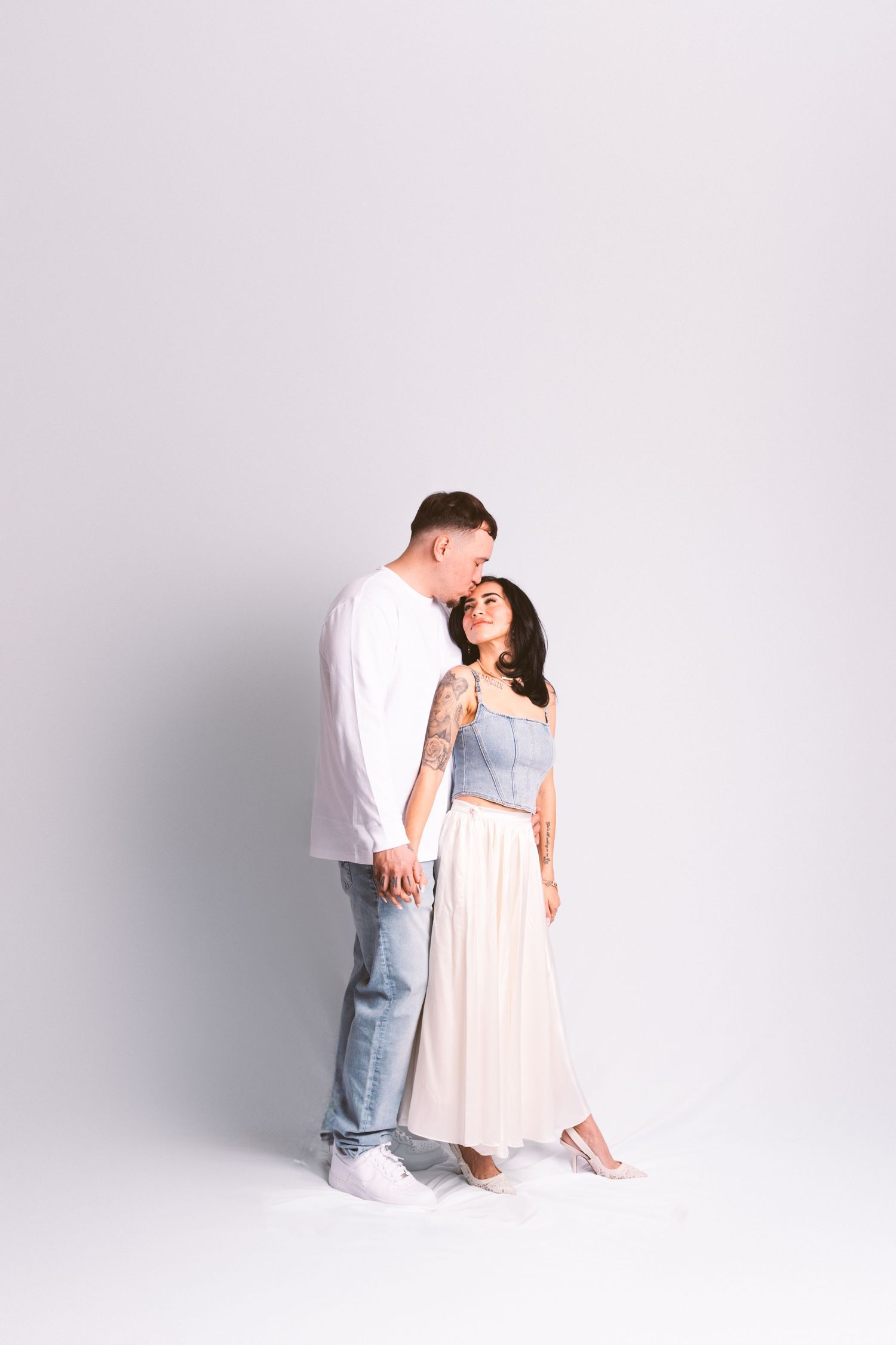 A couple in white and pink outfits share an intimate moment against a light gray studio background.