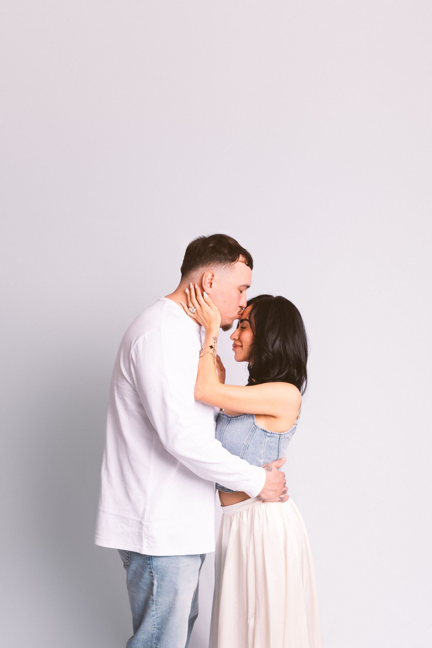 A romantic couple embraces tenderly against a light gray background, dressed in casual white and light colored clothing.