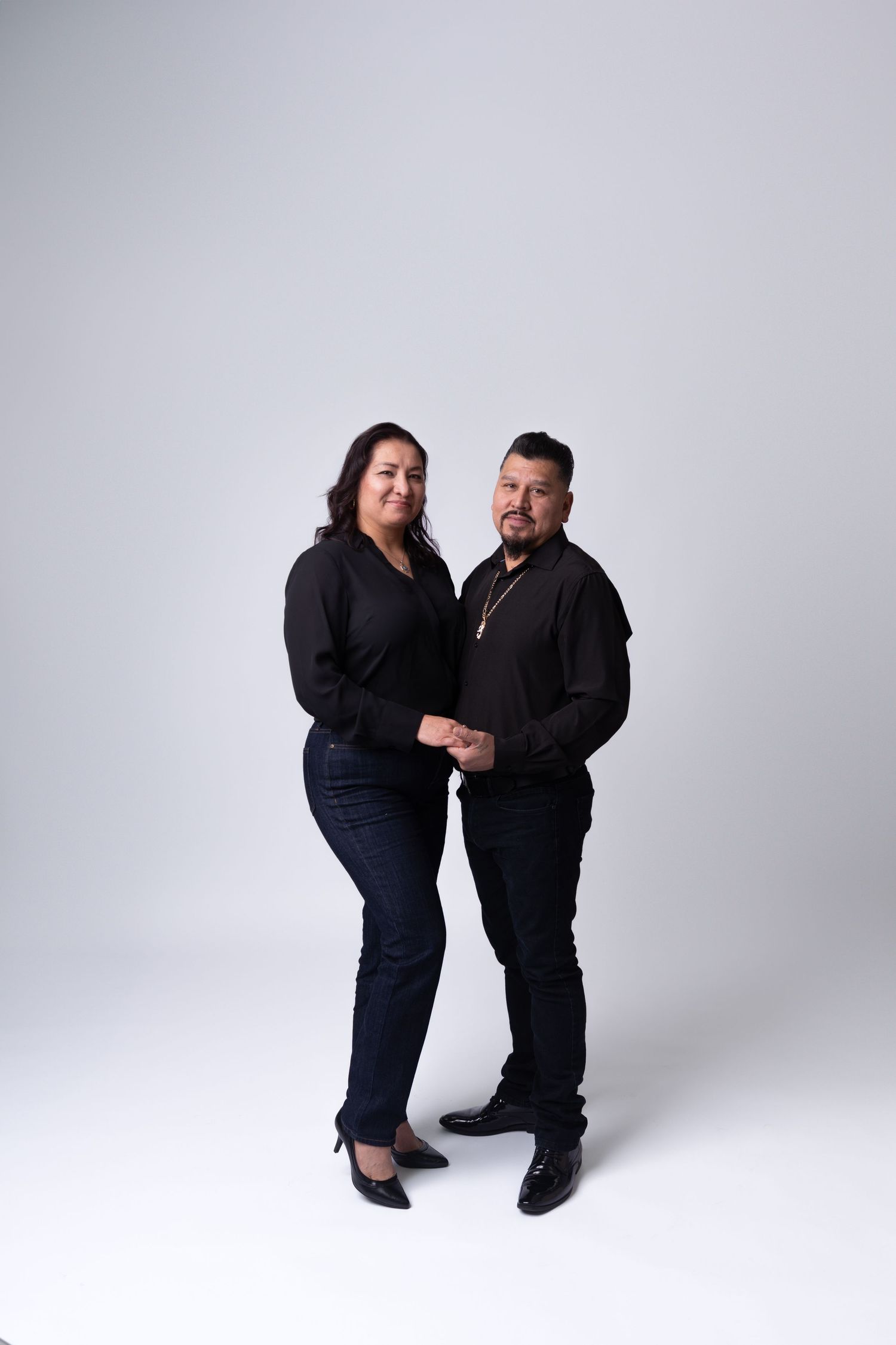 Couple in black outfits poses playfully against white studio background.