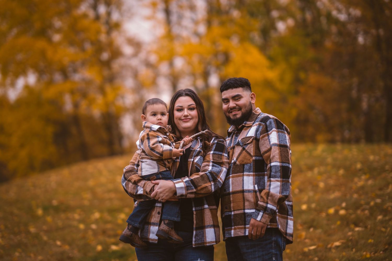 Three people in matching plaid shirts stand together surrounded by fall colors.