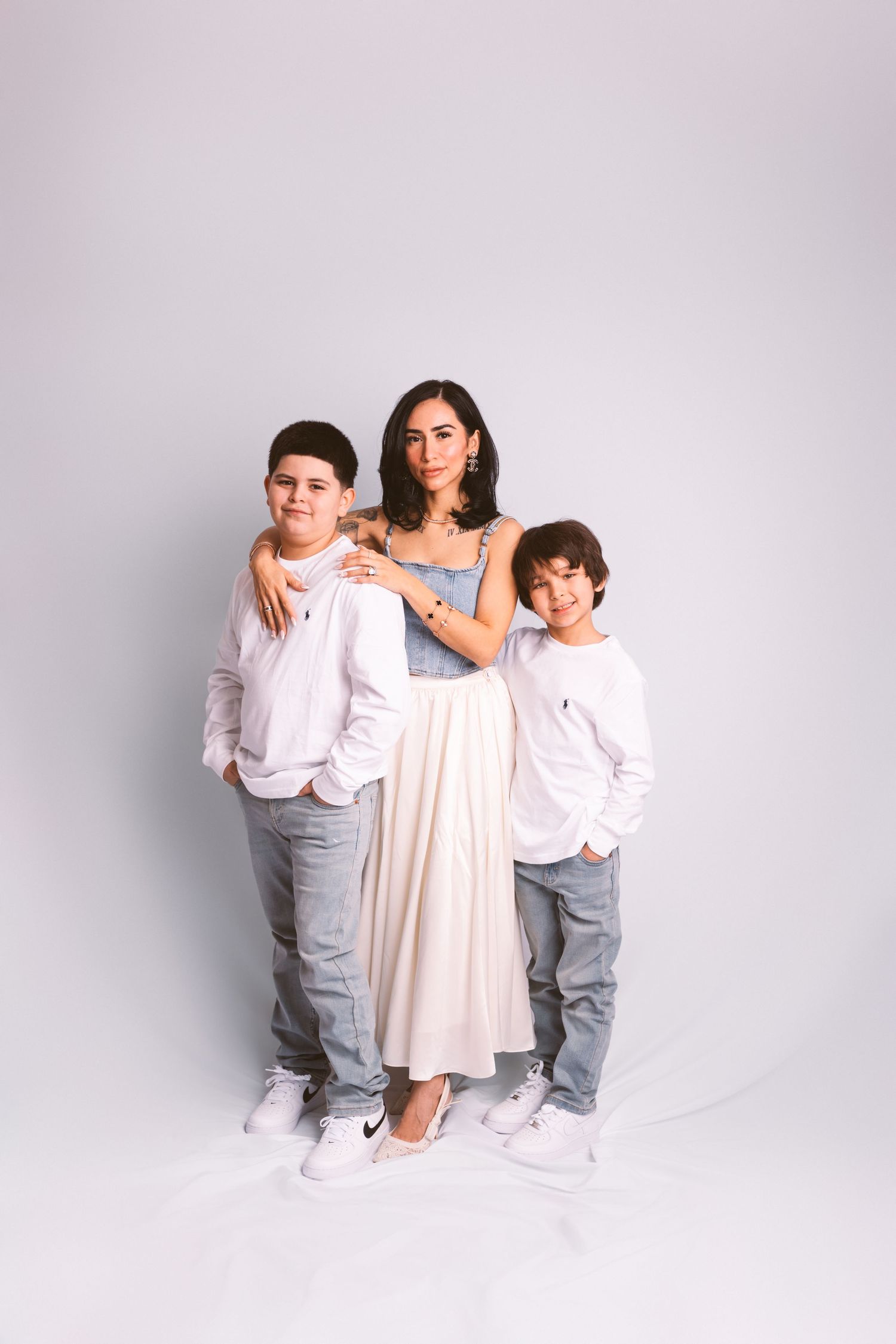 Three young siblings pose together in coordinated white and light pink outfits against a light gray studio backdrop.