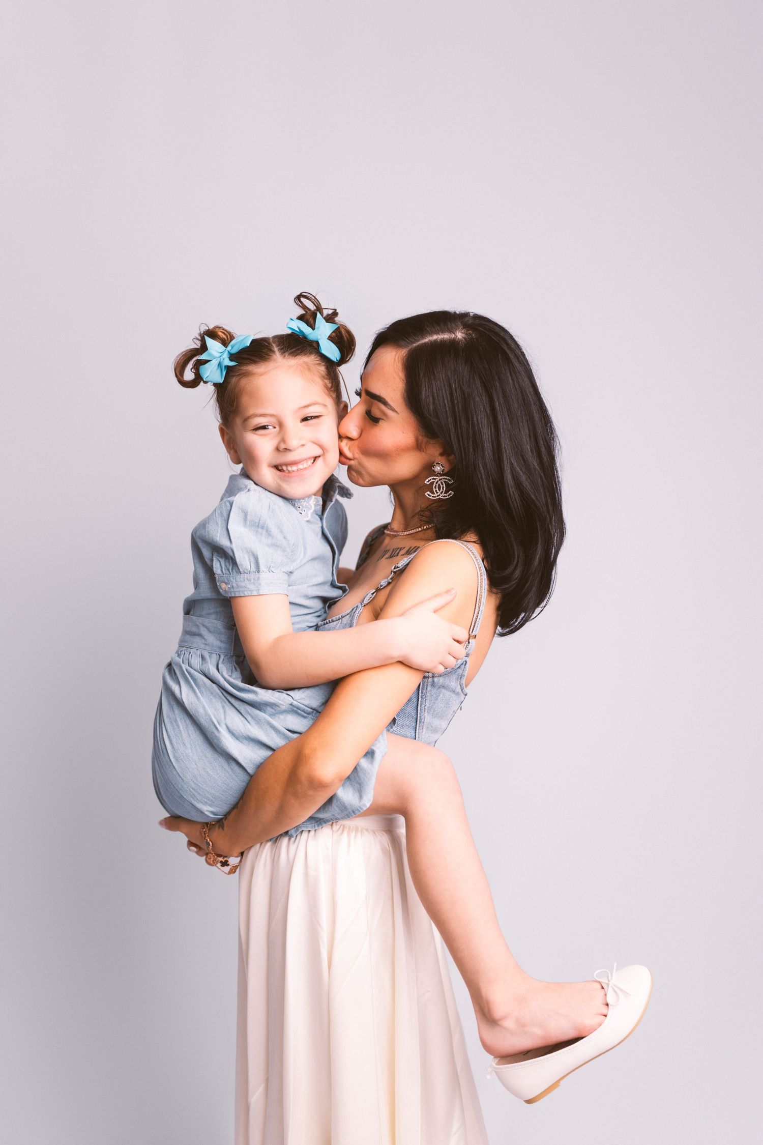 A loving parent lifts and kisses their young child wearing blue against a light gray studio backdrop.