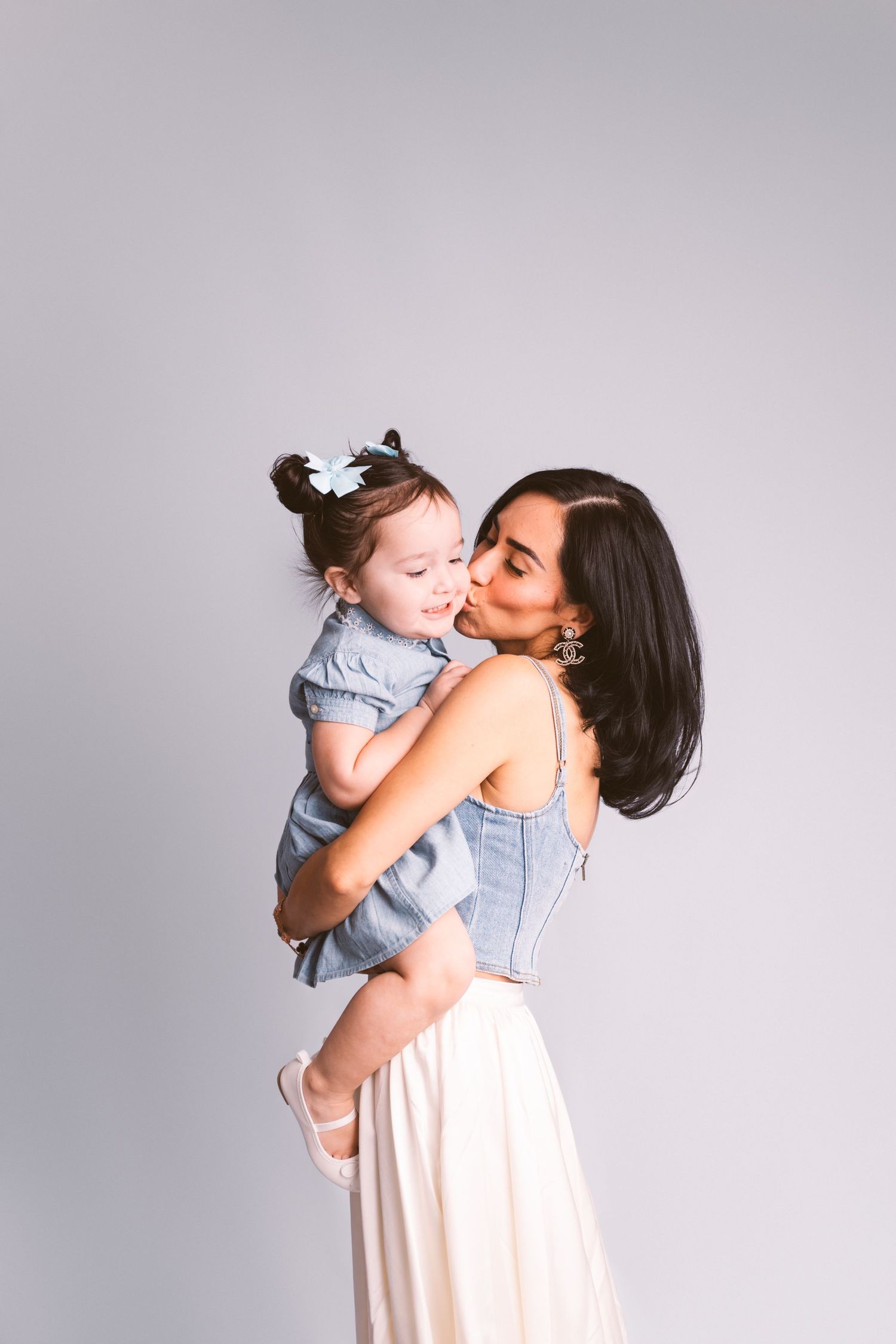A tender moment between parent and child in matching light-colored outfits against a gray studio backdrop.