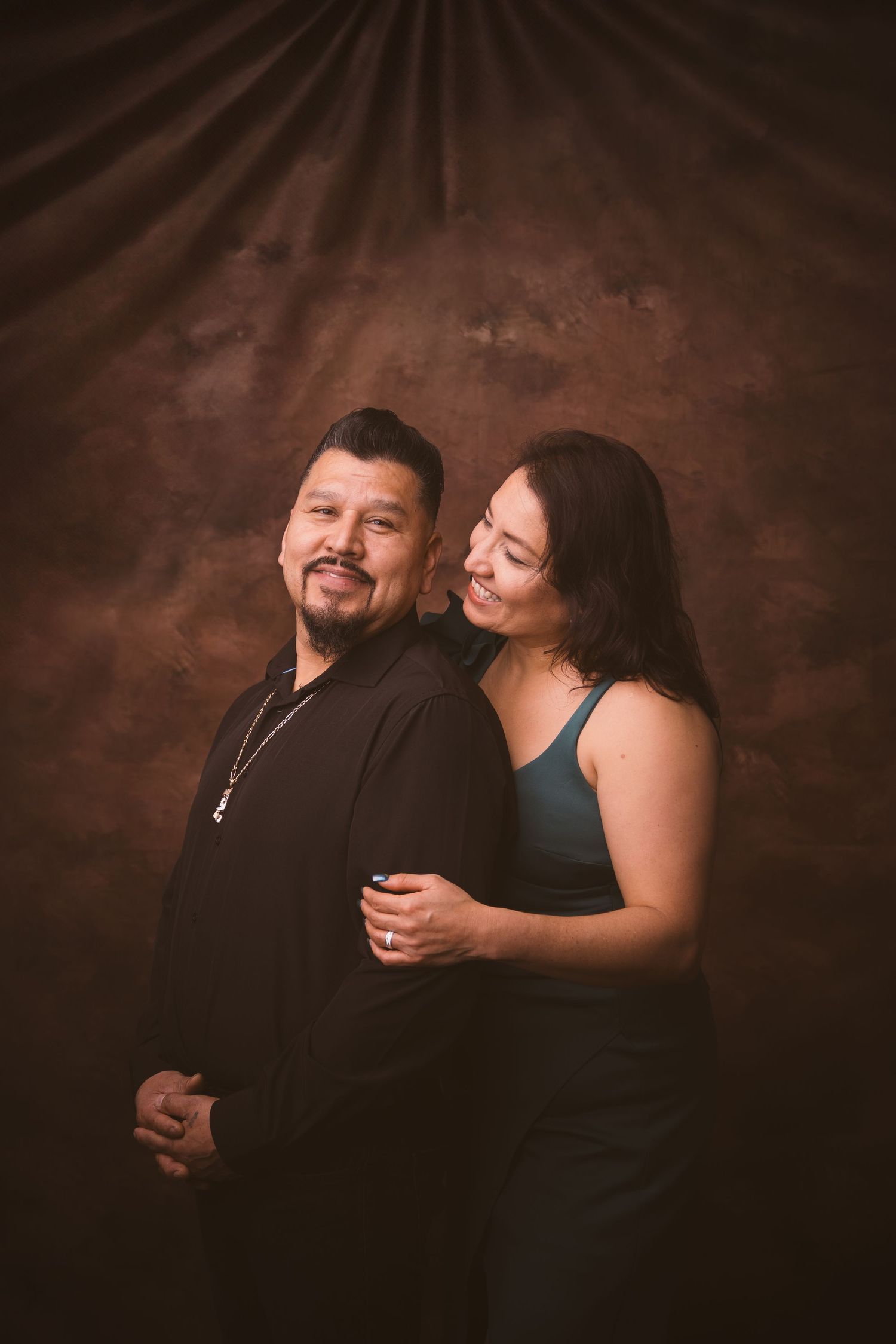 A romantic couple poses together against a dark brown backdrop in an intimate portrait session.