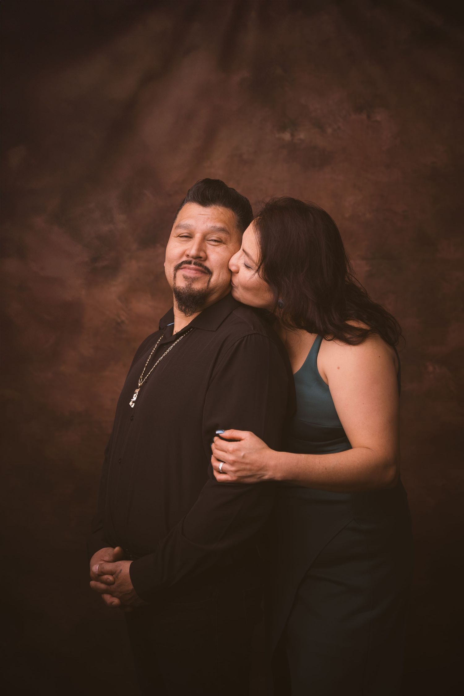 A couple shares an affectionate moment against a dark brown studio background.