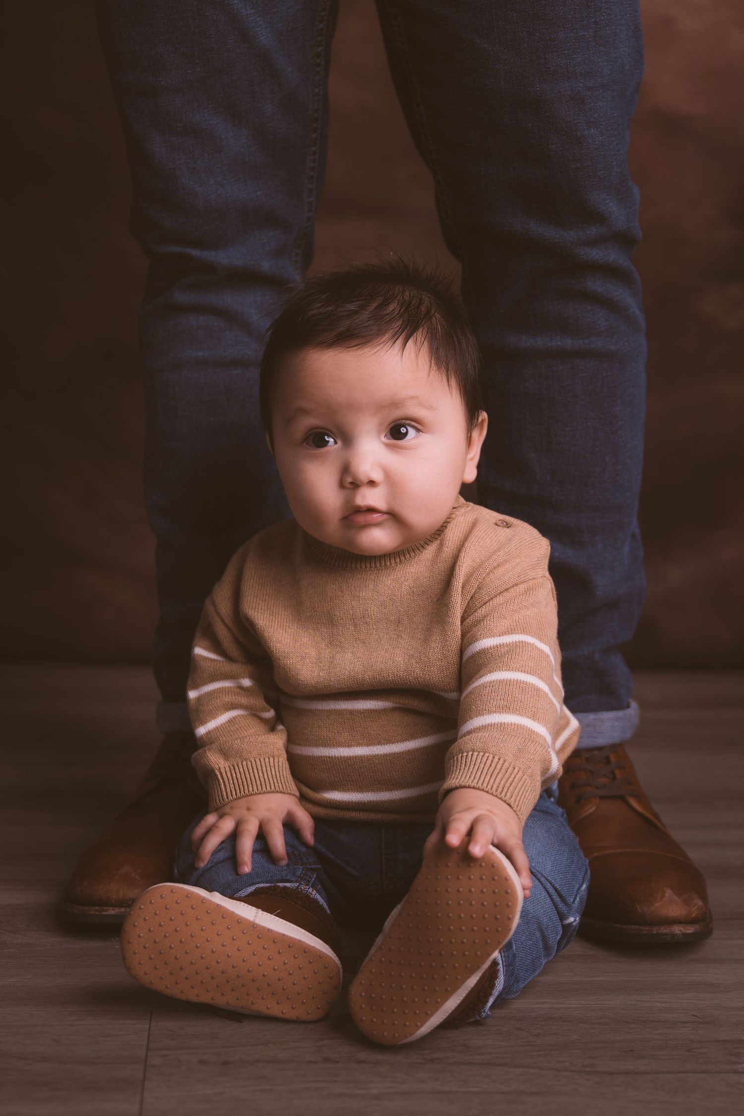 Close-up portrait of baby wearing tan sweater and jeans on dark floor.