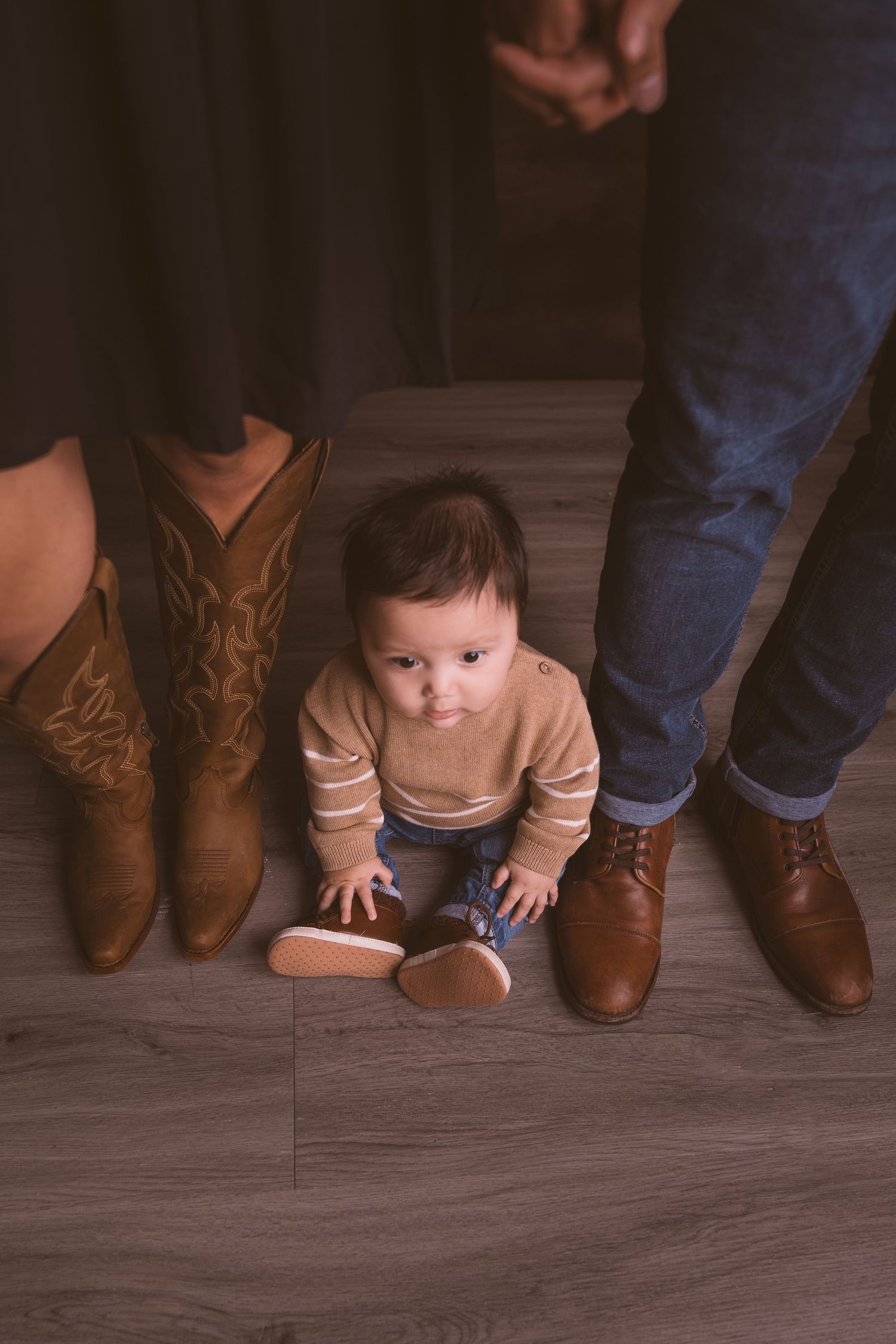 A series of cowboy boots lined up on a wooden floor with a baby crawling between them.