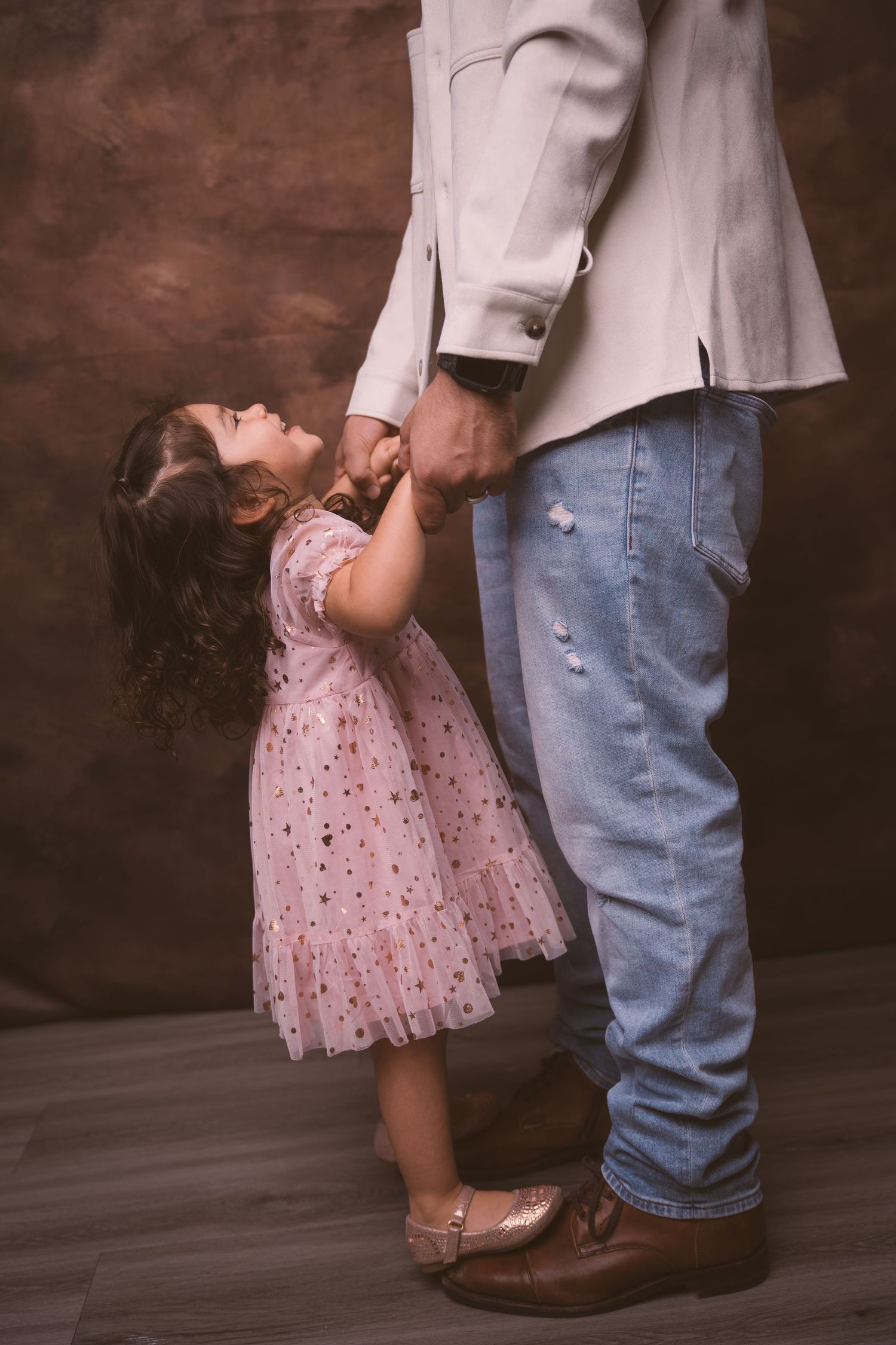 A toddler in a sparkly pink dress stands next to adult legs in a studio setting.