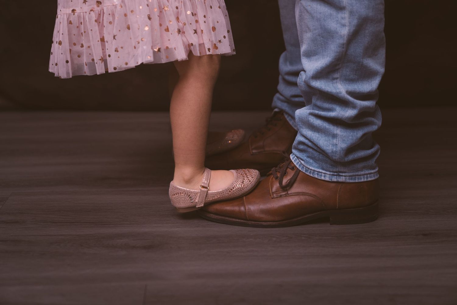 Close-up of contrasting footwear showing brown leather shoes and pink ballet flats on a dark floor.