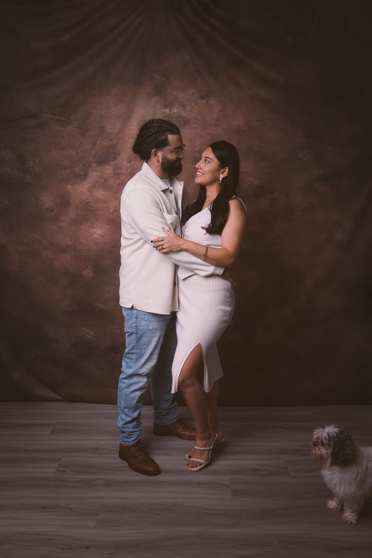 A romantic studio portrait of a couple in coordinated outfits with a small dog in the background.