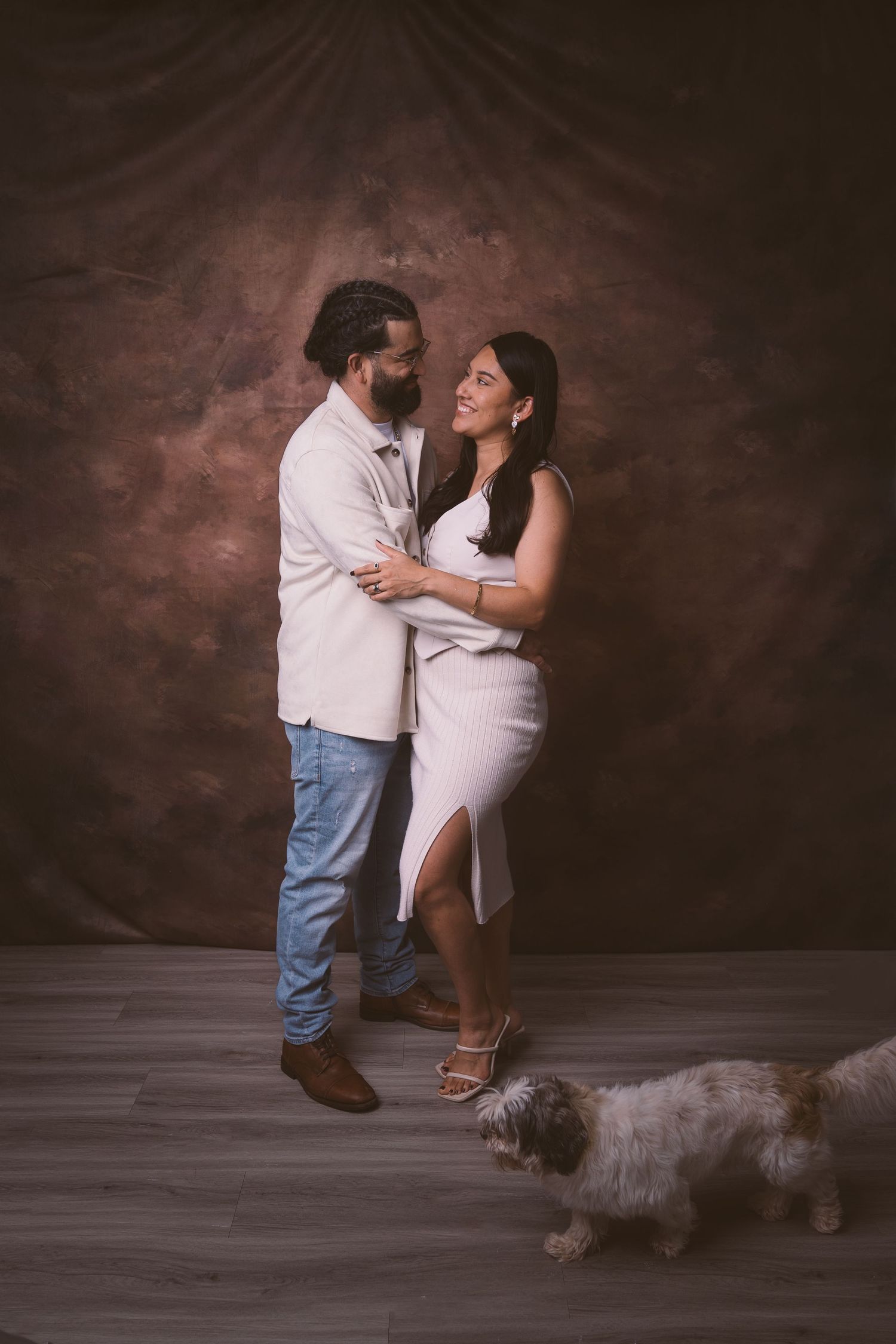 A loving couple poses in a studio with their small dog walking in the background.