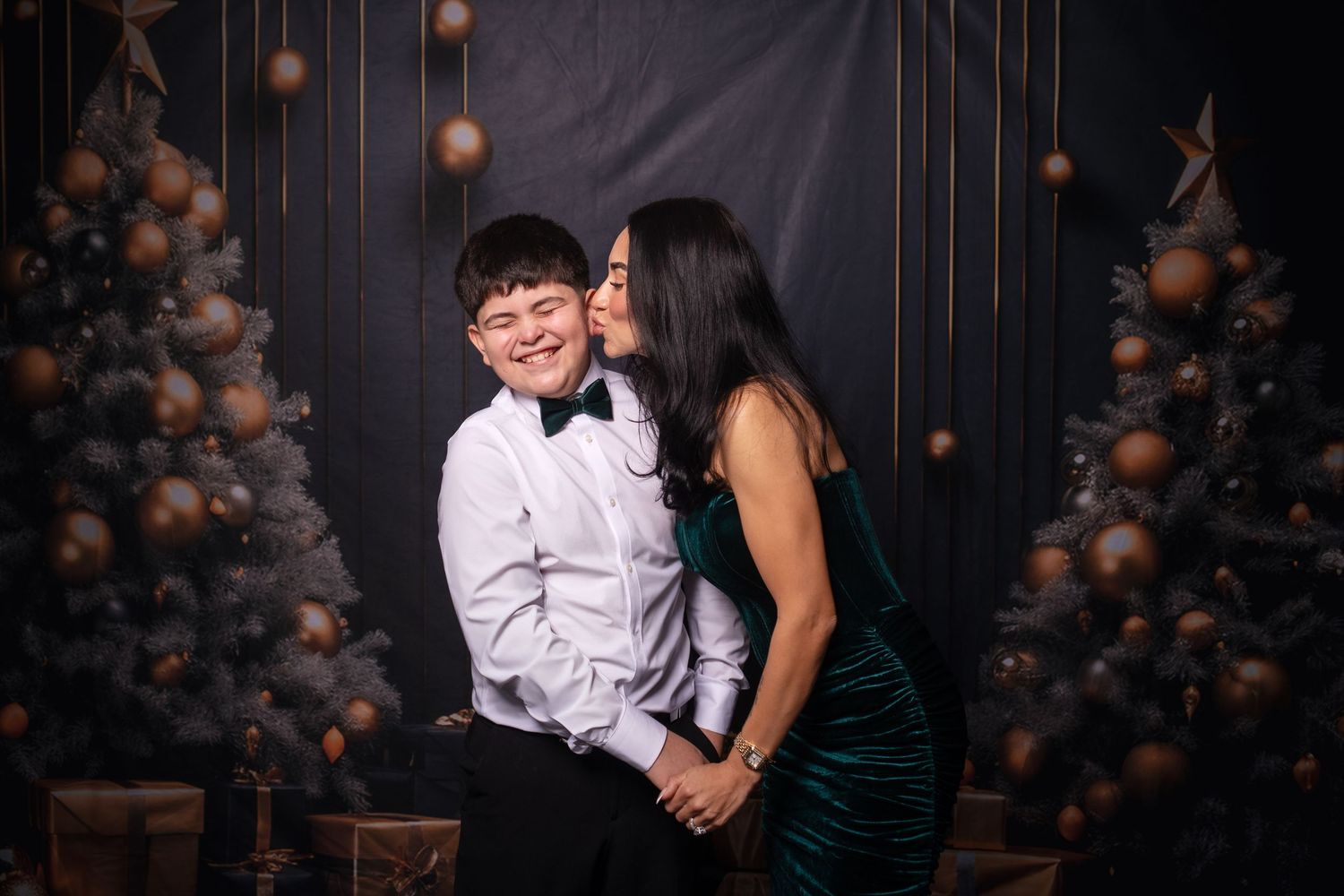 Two people pose together in formal attire against dark holiday-themed background.