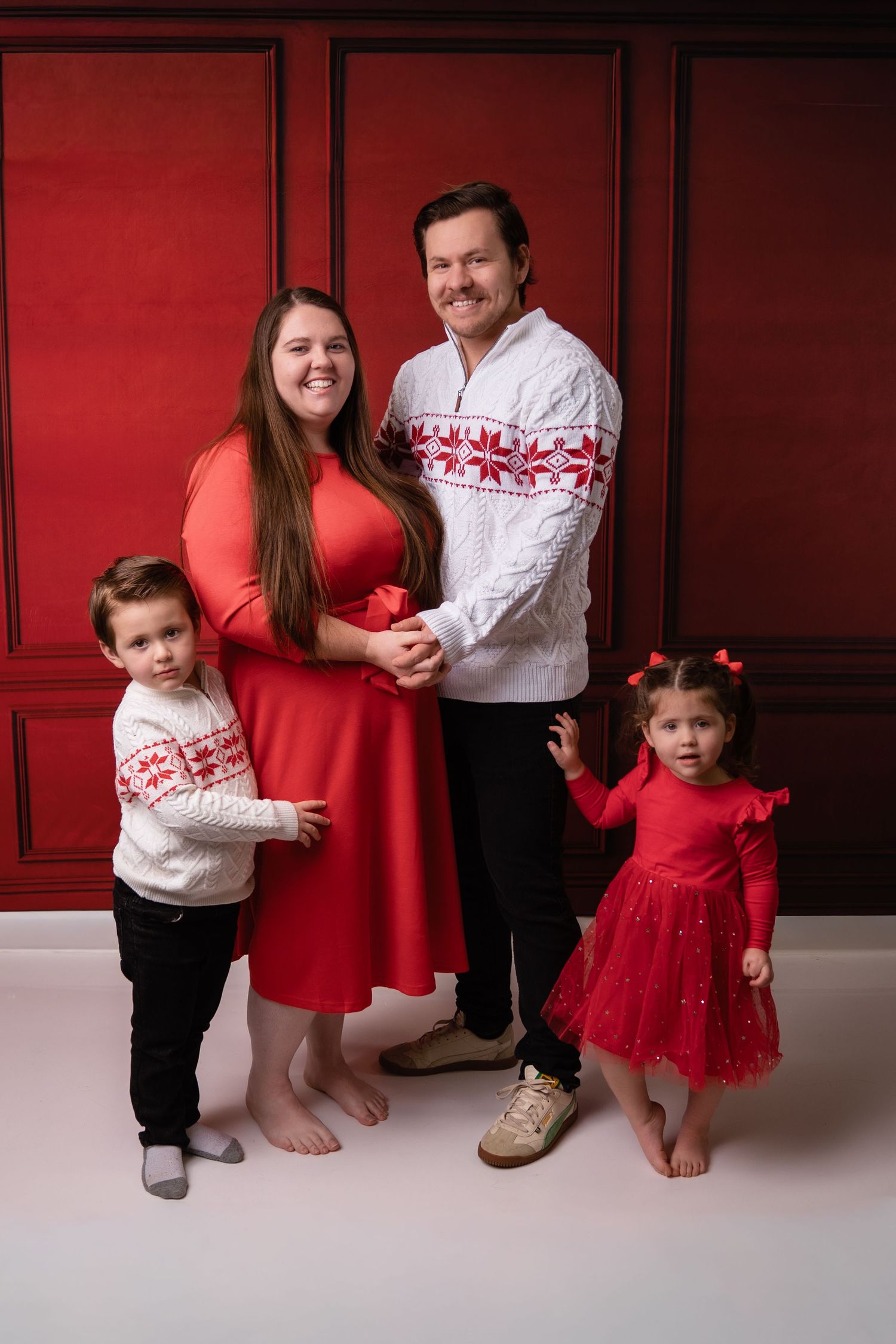 Family holiday portrait with coordinating red and white outfits against red wall backdrop.