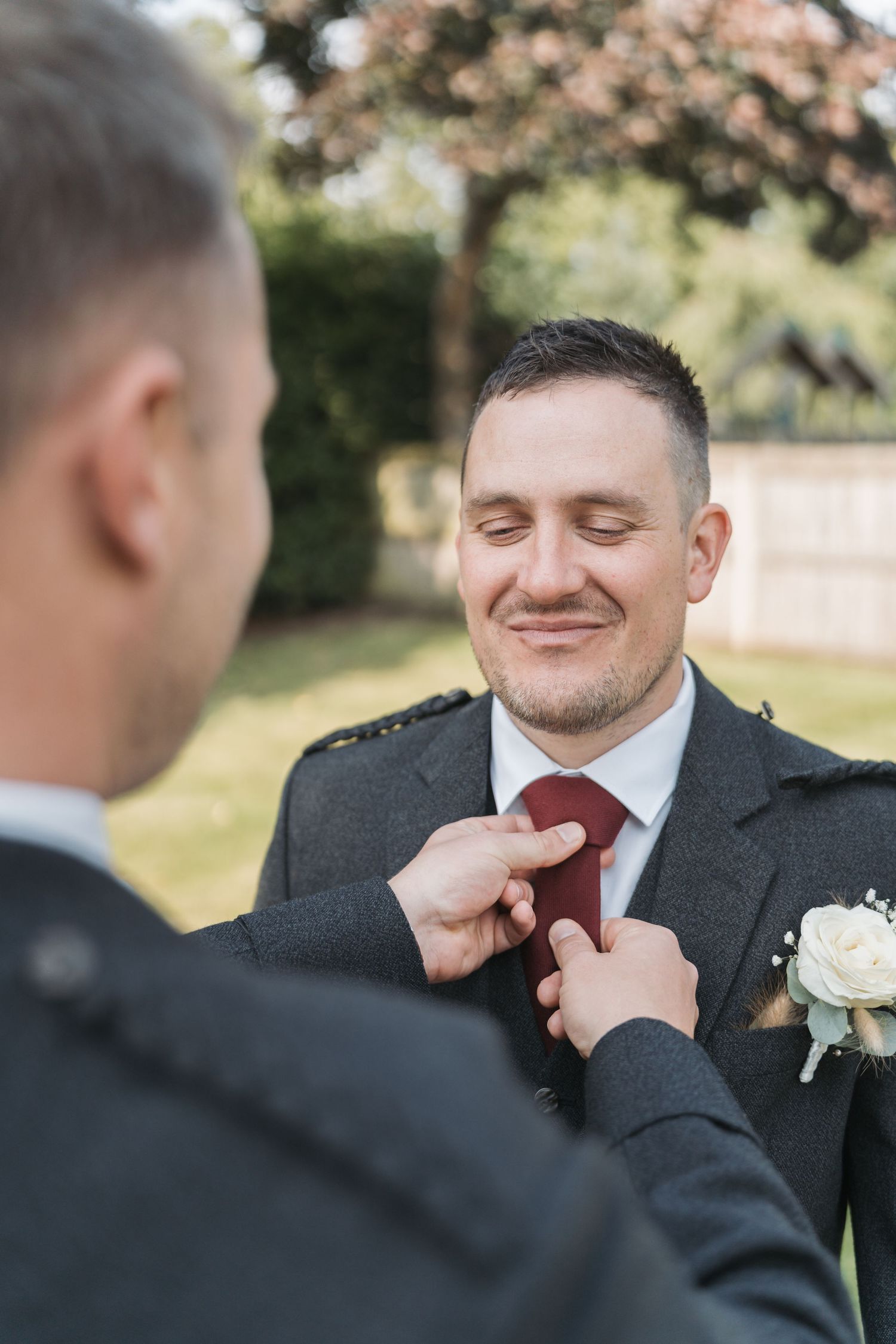 Someone adjusts a red tie and white boutonniere on a smiling person in a dark suit during an outdoor wedding ceremony.