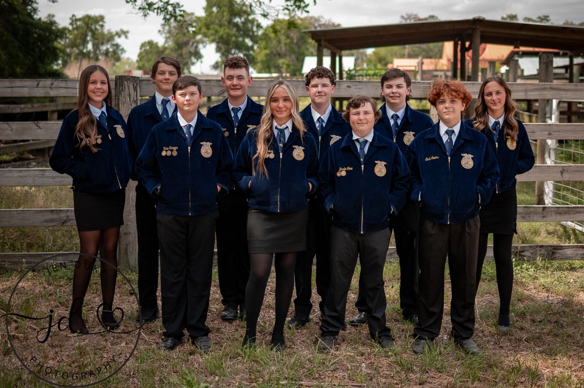 A group in blue FFA jackets gathered outdoors at a rustic wooden fence location for a group photo.