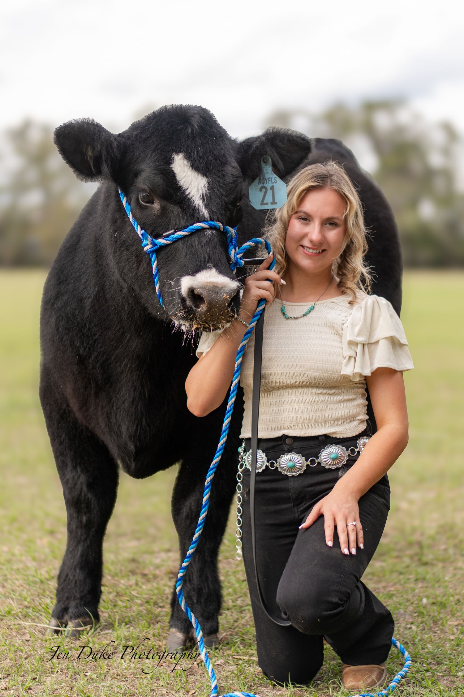 A black horse with blue reins stands in a grassy field with someone next to it during a photo session.
