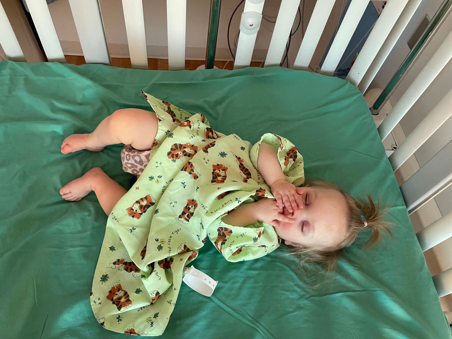 A baby lies in a crib wearing a light green sleeper with teddy bear print on teal bedding.