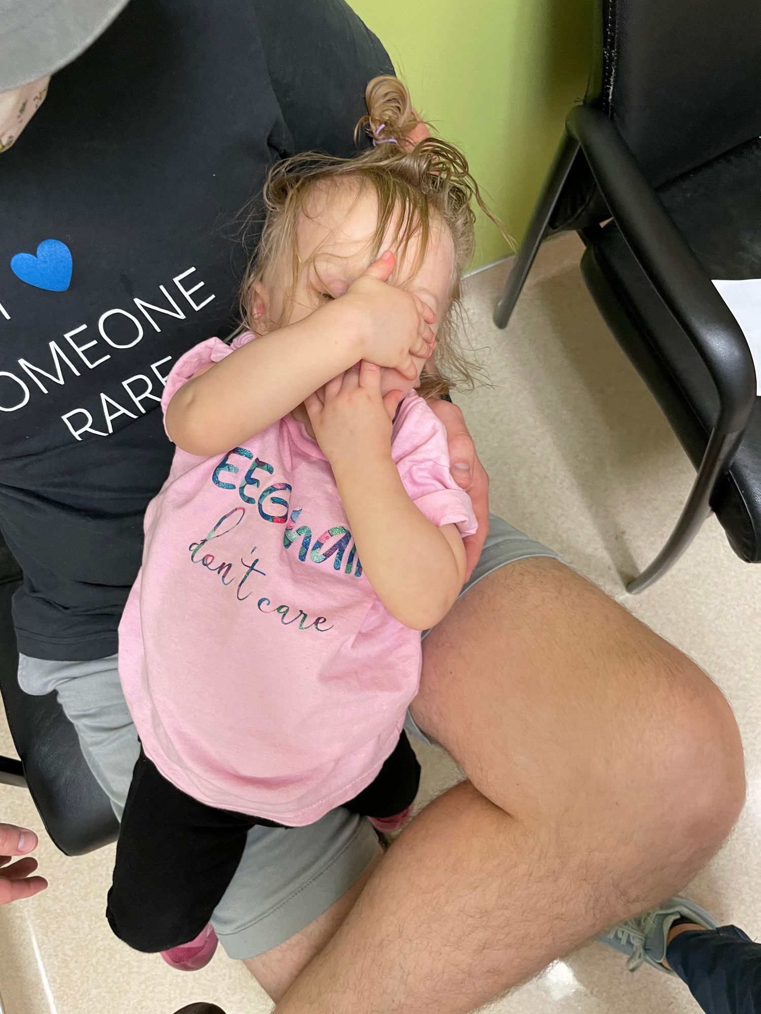 A child in a pink t-shirt sits on someone's lap while covering their face with their hands.