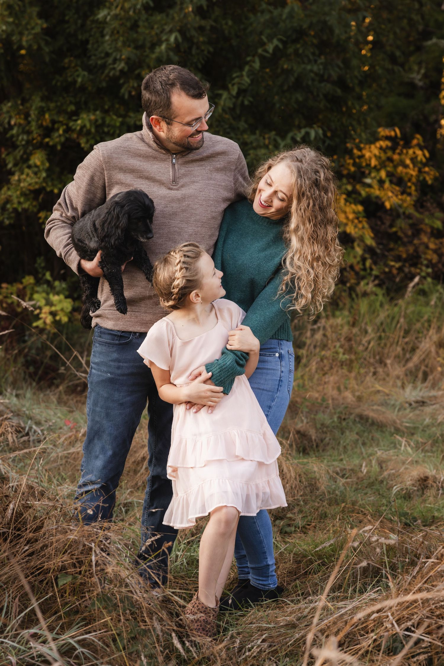 A family shares a loving moment outdoors amongst autumn foliage while holding their small black dog.