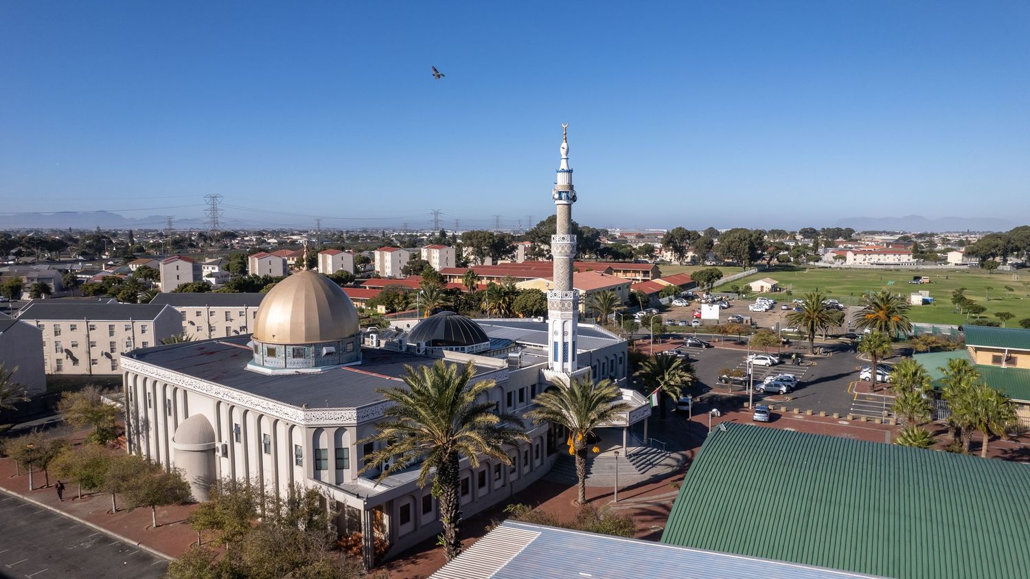 Aerial view of a grand mosque with dome and minaret against clear blue sky in a coastal city.
