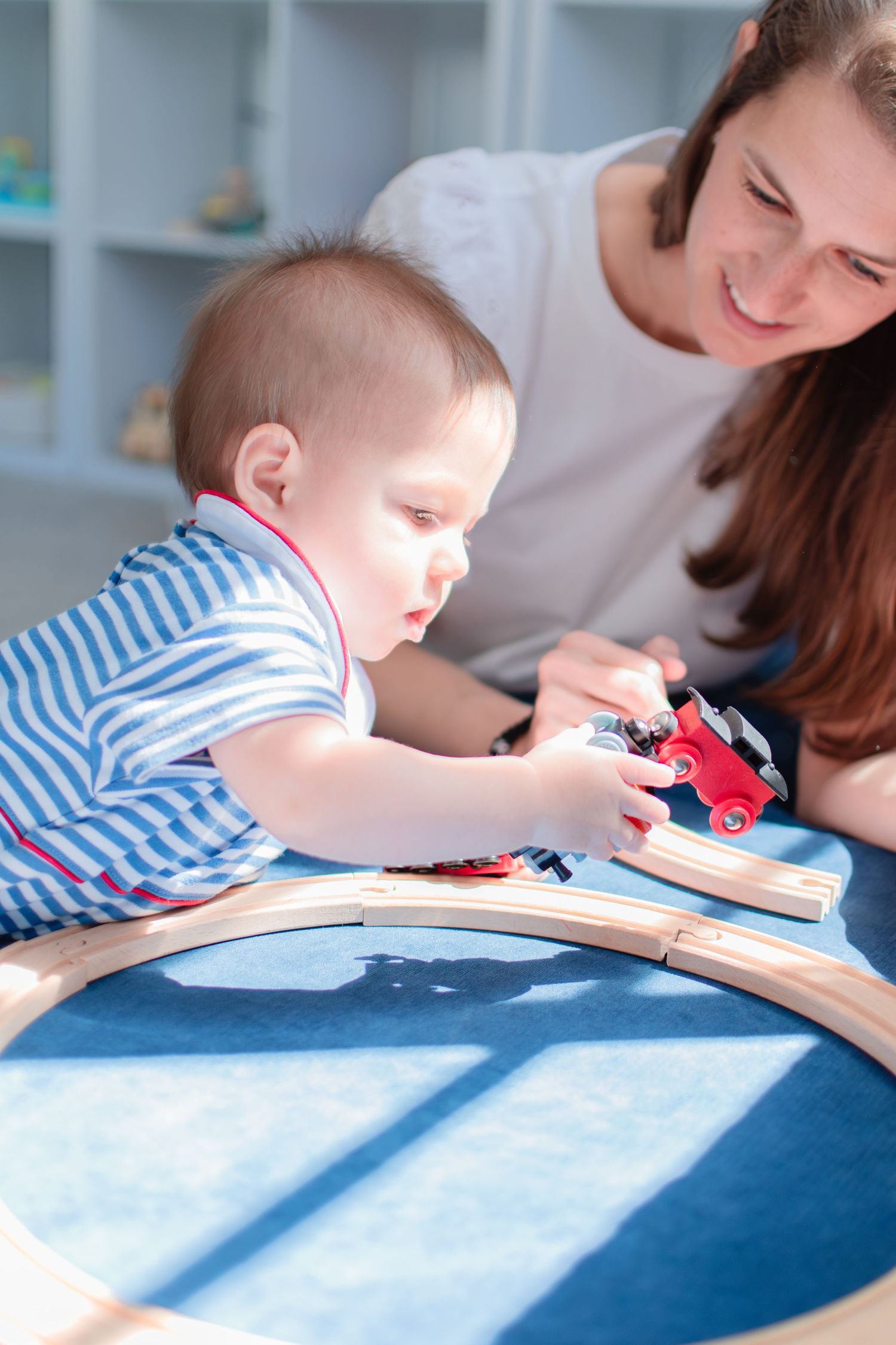 A toddler in striped clothing plays with a wooden toy train set while an adult assists in a bright playroom setting.