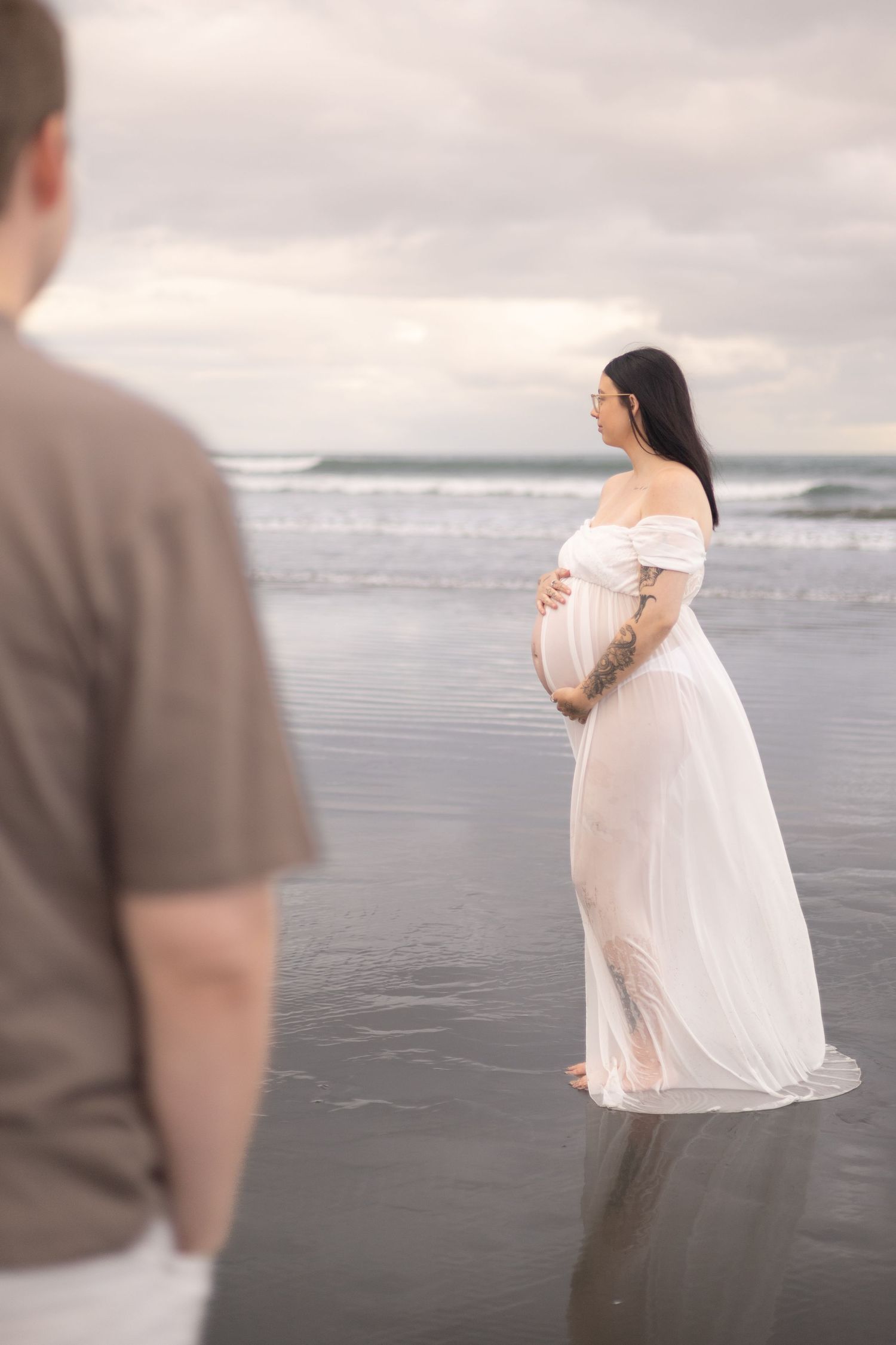 Expecting dad gazing lovingly at his pregnayt partner during a beach maternity photoshoot