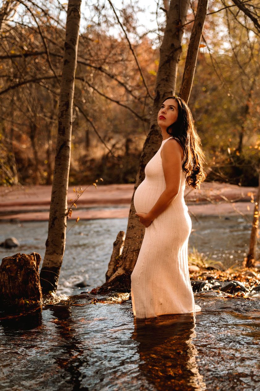 A maternity photoshoot in a stream during autumn featuring a white form-fitting dress against a golden woodland backdrop.