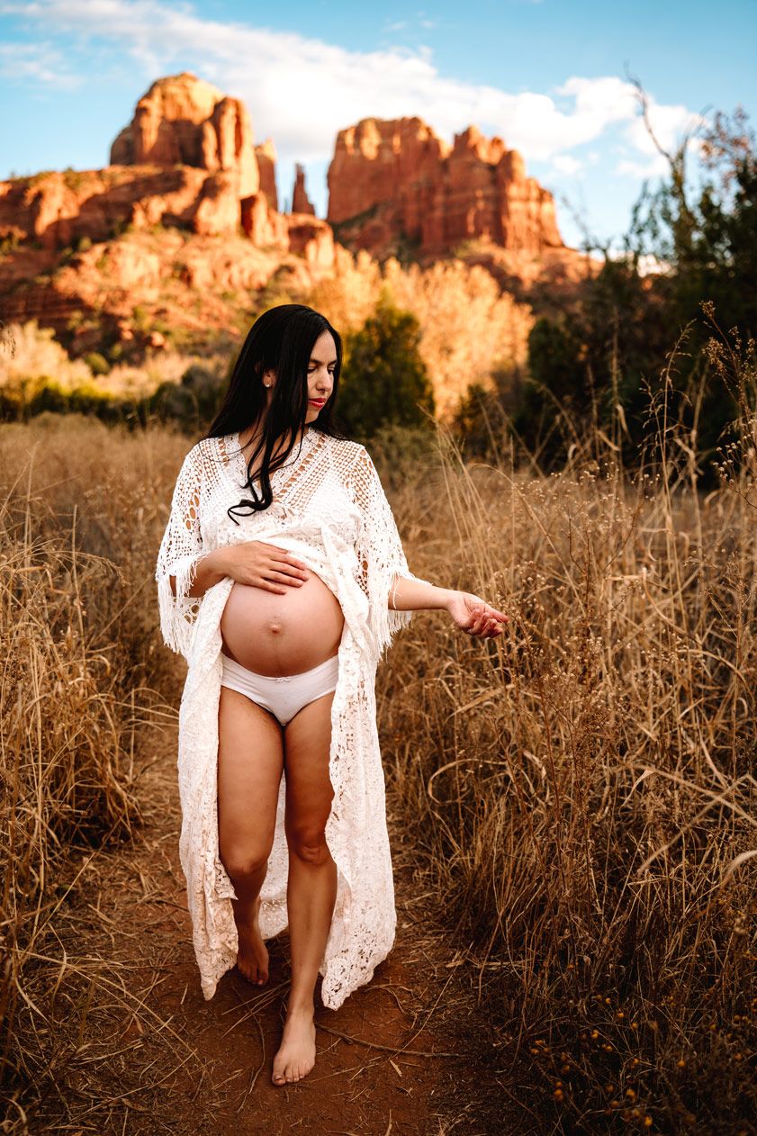 Maternity portrait in lace robe against dramatic Sedona red rock formations and desert brush.