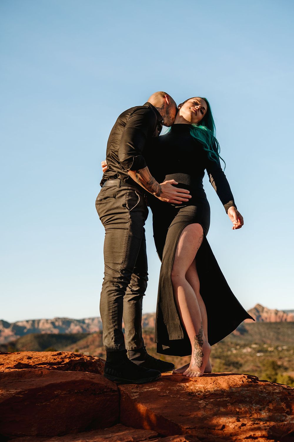 Couple embracing on red rocks in Sedona, woman in black slit dress with teal hair, man kissing her neck at sunset.