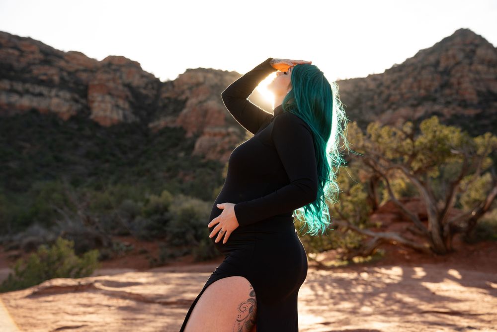 Pregnant woman with teal hair in black dress poses against red rock desert landscape in golden sunlight.