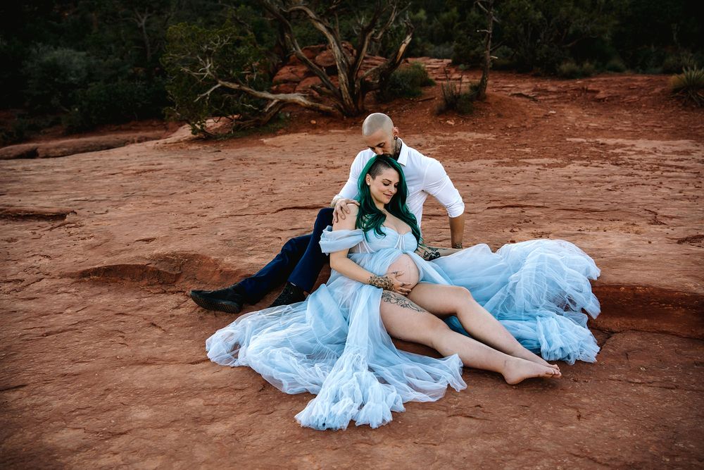 Couple poses romantically on red rock in Sedona, woman in flowing blue gown, man in white shirt, desert landscape backdrop.