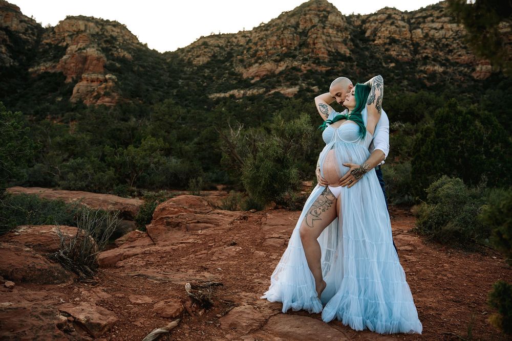 Pregnant couple embracing in Sedona red rocks, woman in flowing blue maternity gown with desert landscape backdrop.
