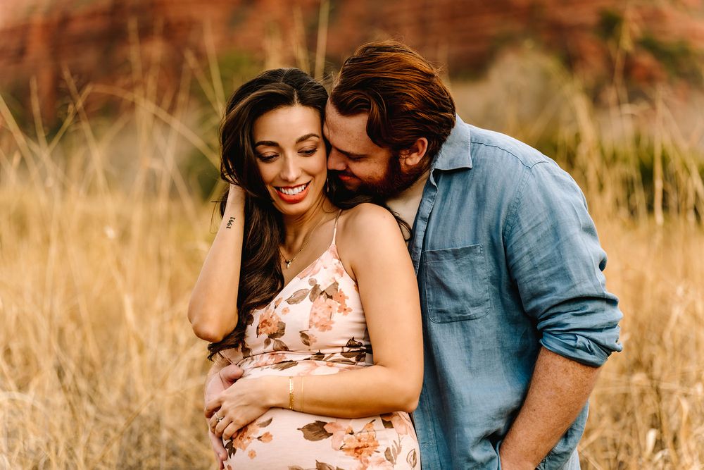 A couple shares a tender moment in a golden field, the man kissing a smiling pregnant woman in a floral dress.