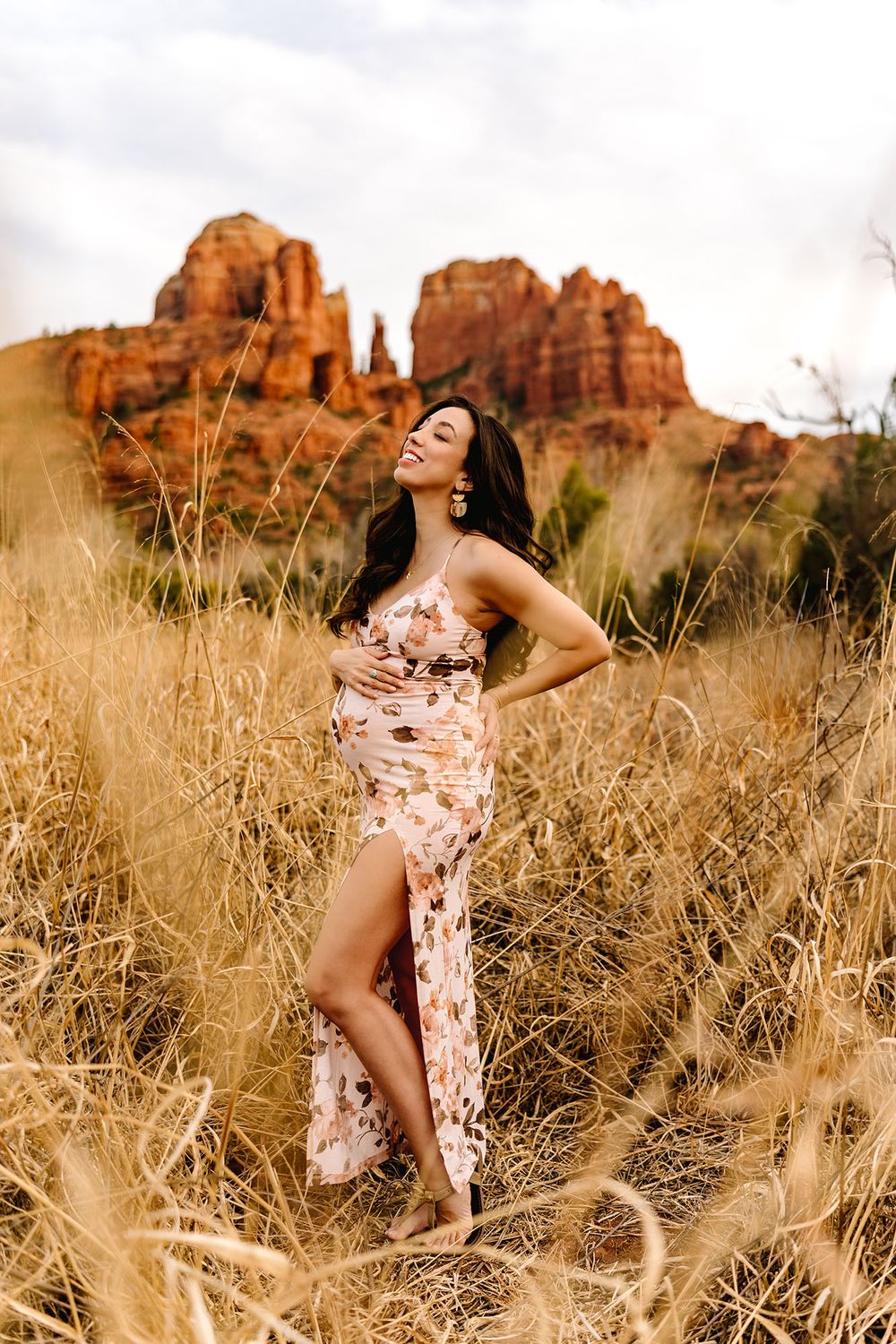 Smiling pregnant woman in floral dress standing in tall golden grass with Sedona red rocks.