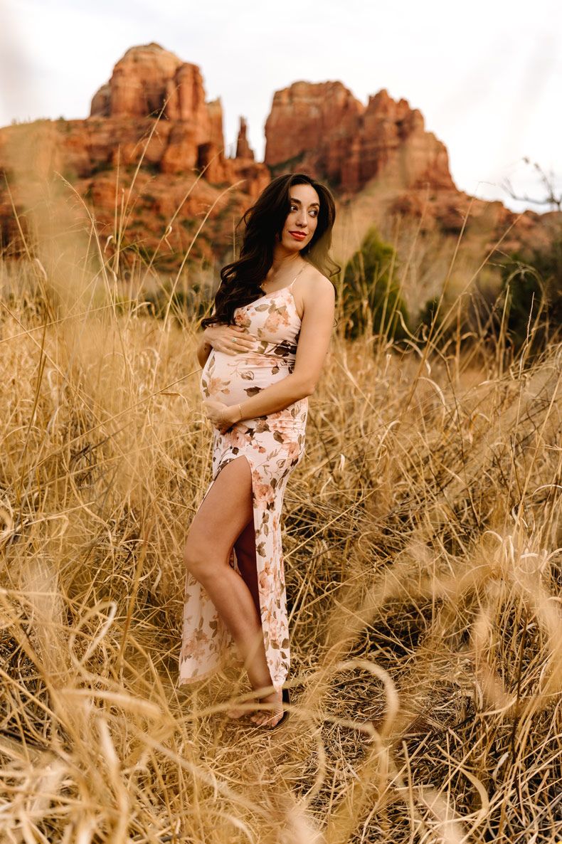 A maternity portrait in a floral dress among desert grasses with Sedona red rocks in the distance.