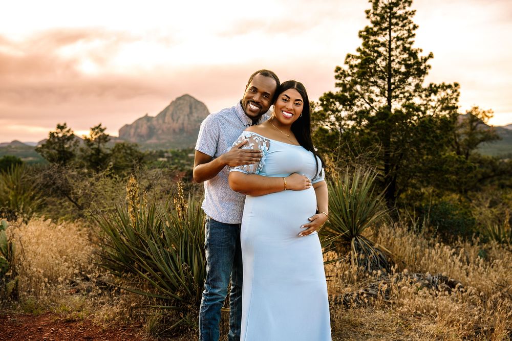 Couple poses in desert landscape at sunset, pregnant woman in blue gown with mountains and pine trees behind them.