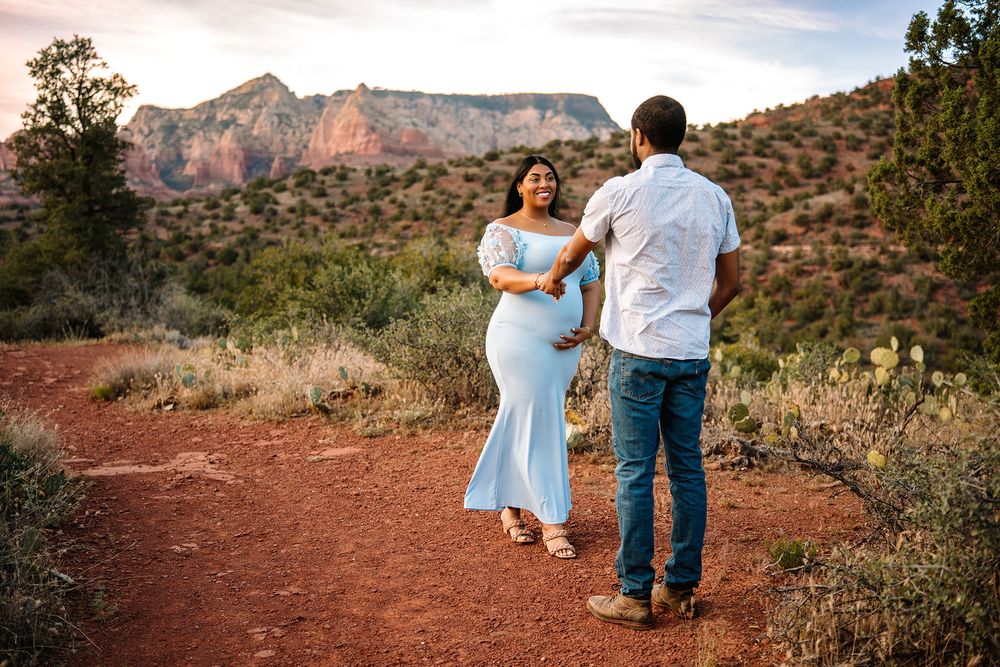 Pregnant couple dancing on red dirt trail in Sedona Arizona with stunning red rock mountains at sunset.