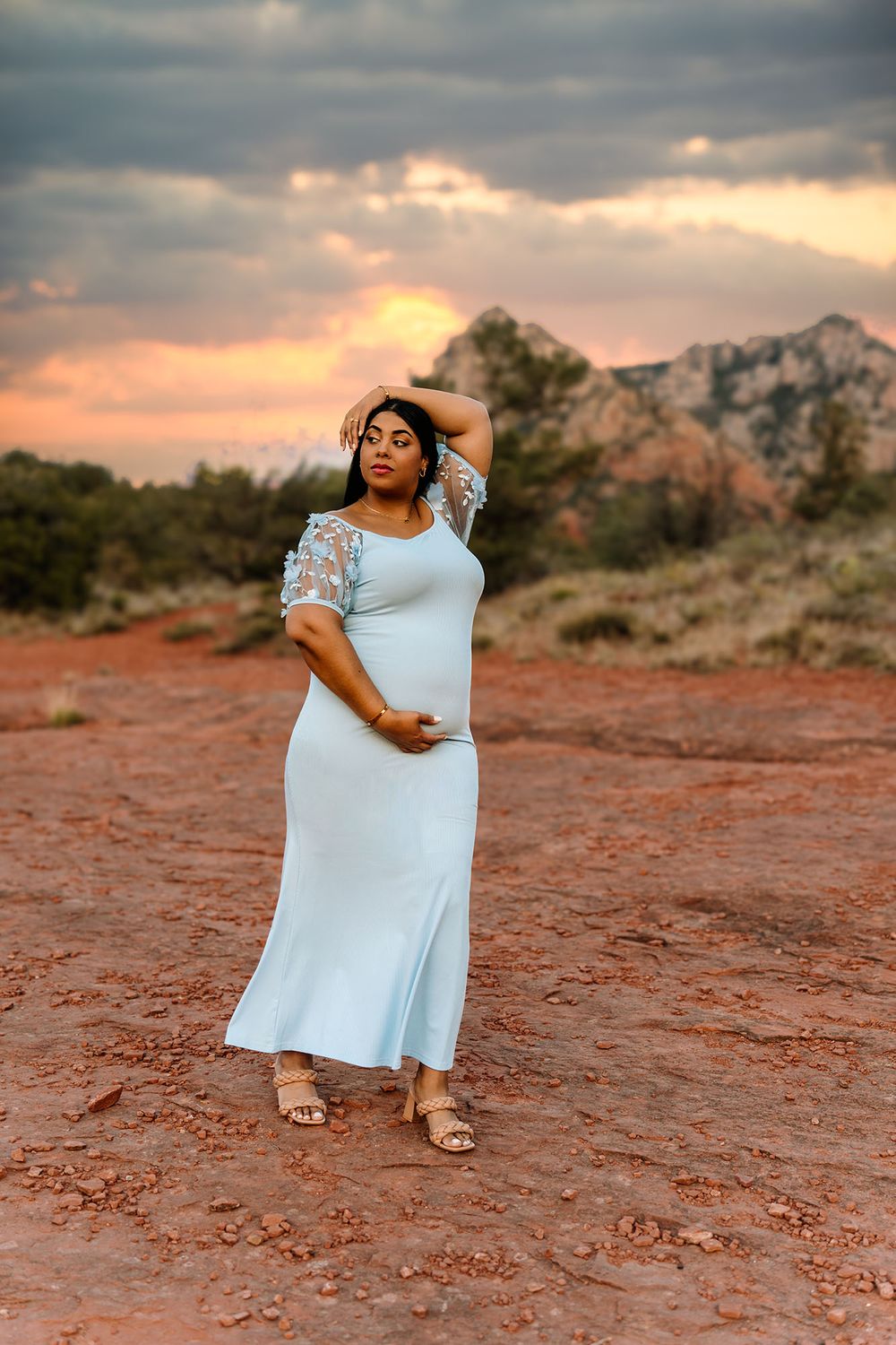 Woman in light blue maxi dress poses in Sedona red rock desert landscape at dramatic sunset.