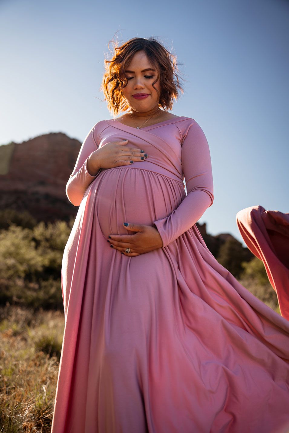 A flowing pink maternity dress catches light against desert cliffs.