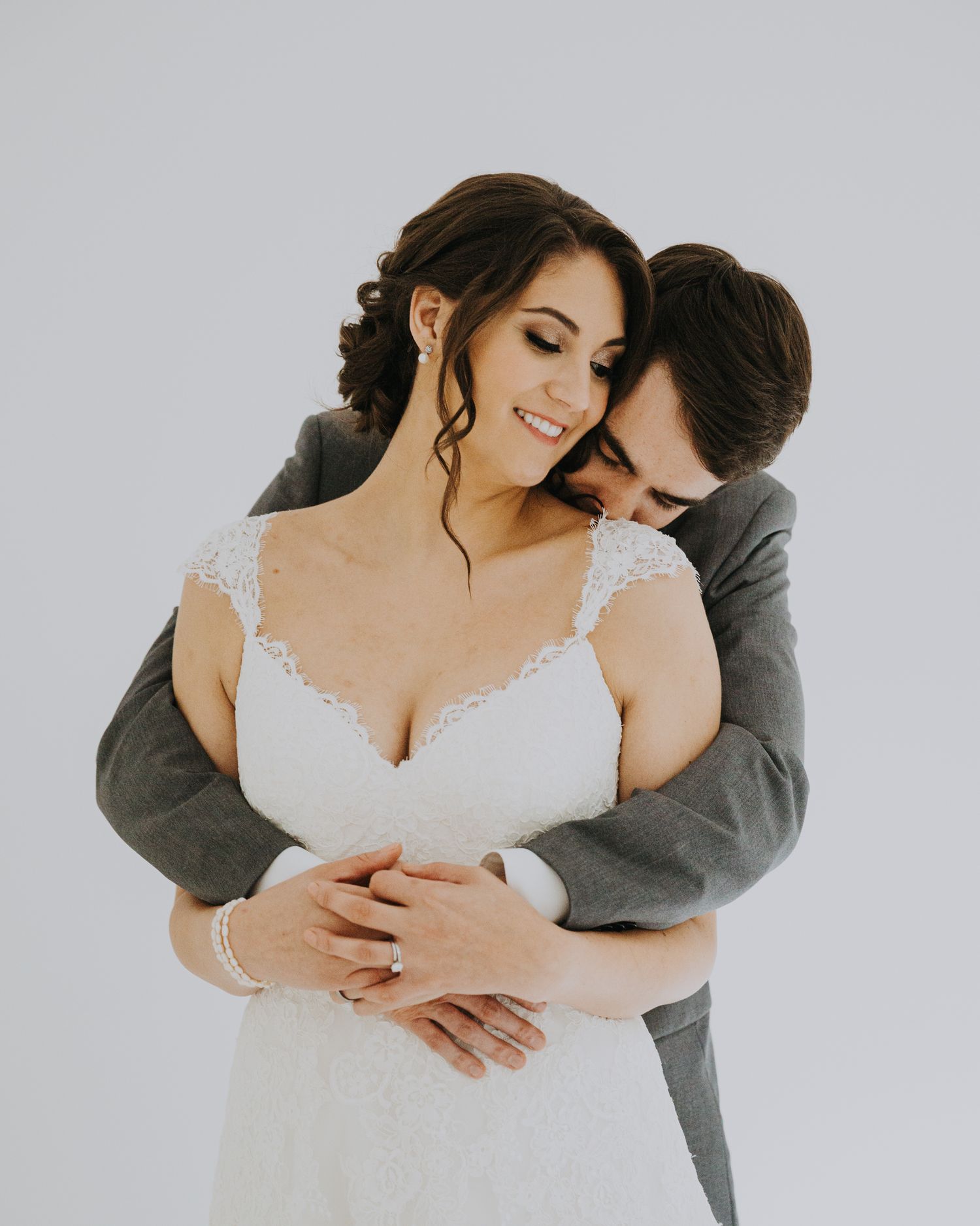 A couple in wedding attire embrace intimately against a white background, with the groom hugging the bride from behind.
