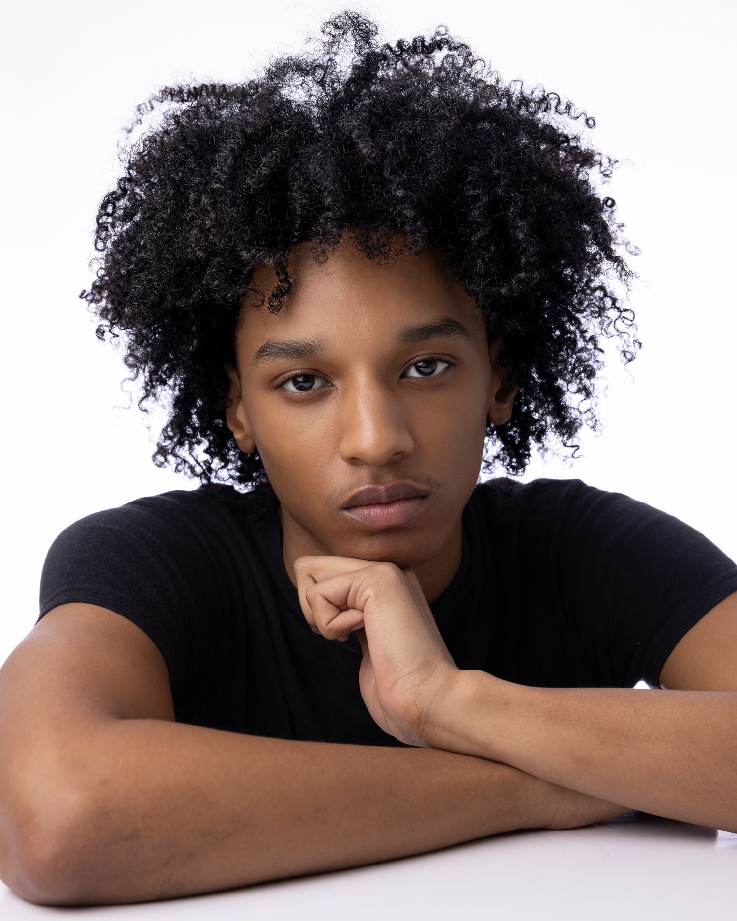 Person with natural curly afro hair wearing black t-shirt poses against white background with hand under chin.
