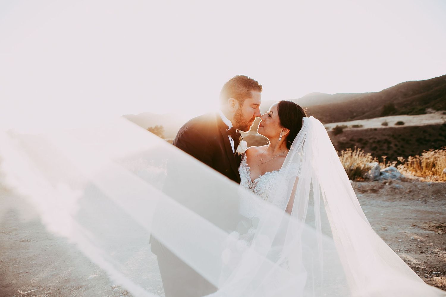 Bride and groom at their orange county wedding with veil flowing and the sun setting in the background as they embrace.